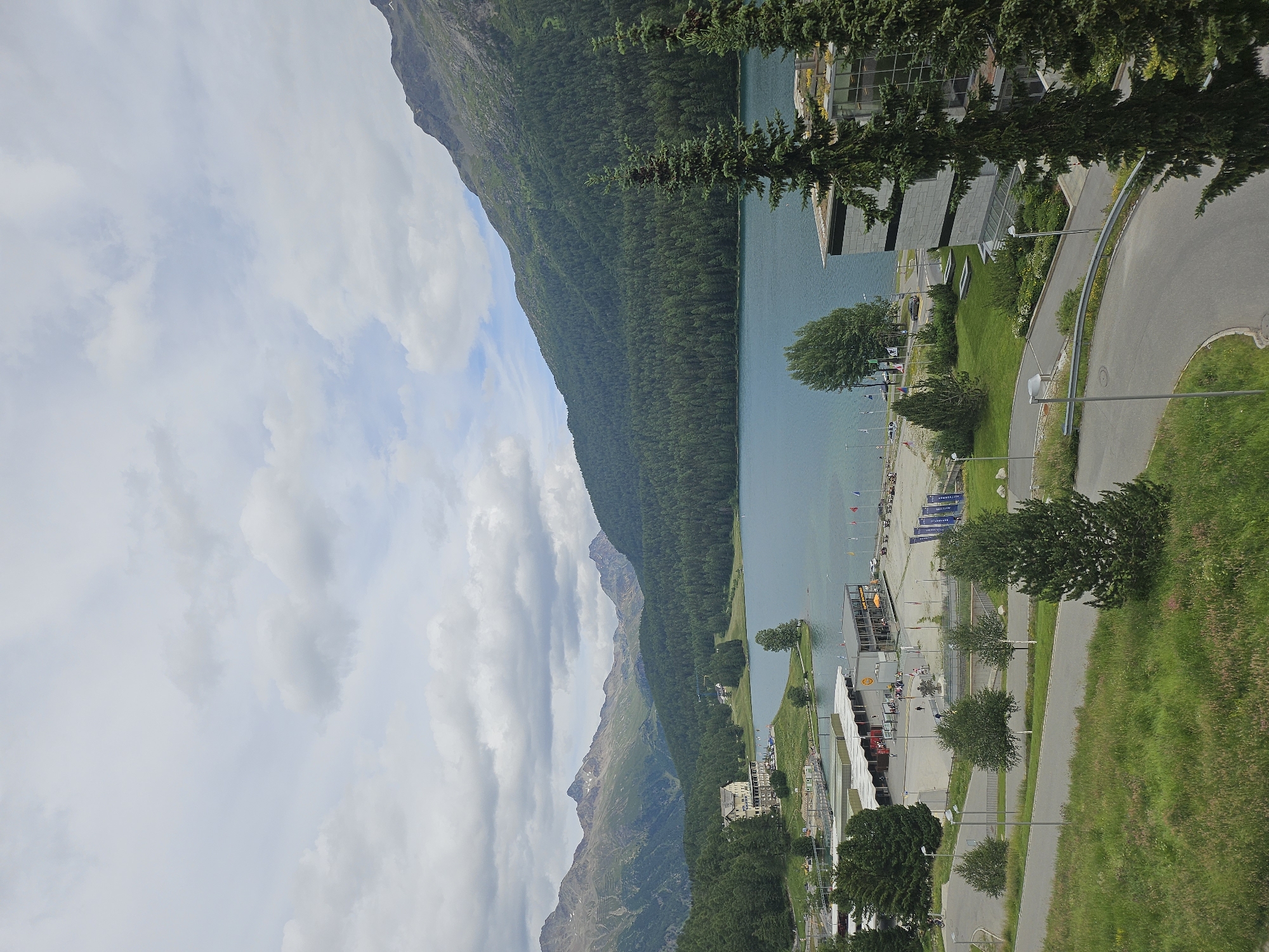 An elevated, wide-angle, landscape-orientation, daytime, eye-level shot captures a serene view of the St. Moritz lake in St. Moritz, Switzerland. The tranquil, blue lake dominates the center of the frame, bordered by a dense, dark green forest that blankets the foothills of majestic, rugged mountains in the background. The sky above is a soft, cloudy gray, with patches of blue peeking through.
Along the lakeshore, a resort-like setting unfolds. A large, light-colored hotel with multiple windows and a red-tiled roof is prominently situated on a grassy incline. Adjacent to the hotel, a series of modern, flat-roofed buildings with glass facades and stone exteriors line the water's edge. These structures appear to house businesses or recreational facilities, with several people visible on patios and walkways. Boats are docked in a marina area, and flags flutter in the breeze.
In the foreground, a paved road with a metal guardrail curves through a lush, green landscape dotted with trees and grass. Several streetlights are visible along the road. The overall impression is one of a peaceful, picturesque mountain resort town.