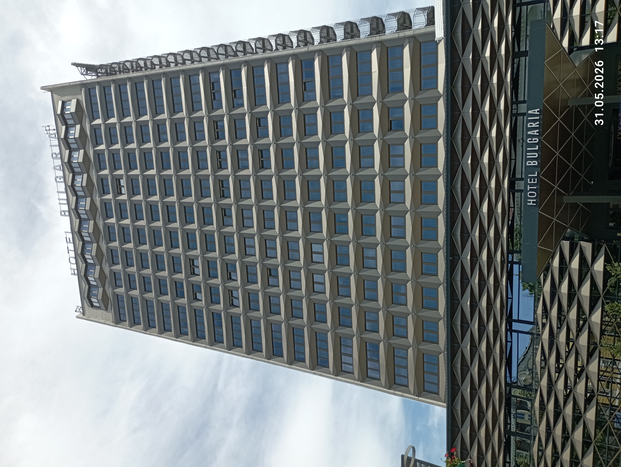The image shows the Hotel Bulgaria in Burgas, Bulgaria. The building is a tall, rectangular structure with many windows arranged in a grid-like pattern, creating a geometric facade. A structure with a geometric diamond pattern is adjacent to the building. The hotel's name is visible on the building itself. The image has a timestamp of "31.05.2026 13:17". The sky is visible in the background, with scattered clouds.