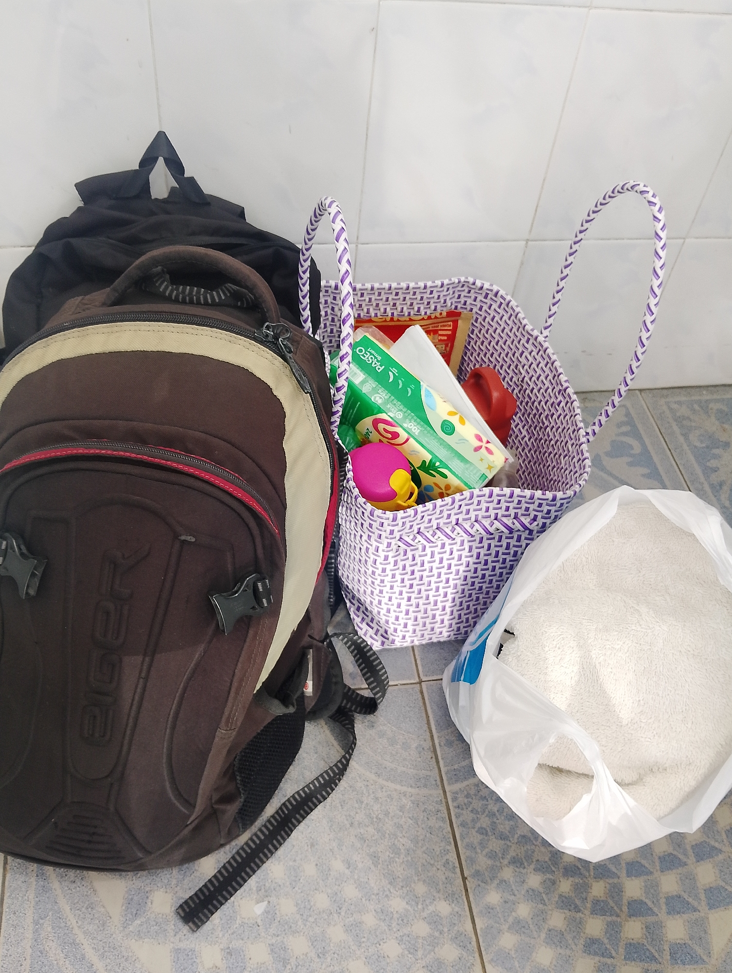 The image displays a collection of items on a tiled floor in what appears to be an indoor setting, possibly a covered porch or hallway, given the visible wall tiles. The items include a brown and tan backpack with straps, a black duffel bag, a woven purple and white basket containing a package of tissues, a red object, and a pink and yellow toy, and a white towel inside a clear plastic bag. The backpack has the brand name "Eiger" embossed on its front. The overall scene suggests preparation for a trip or the storage of personal belongings. The context of "City of Pematang Siantar, Indonesia" is not visually discernible from the provided image itself.