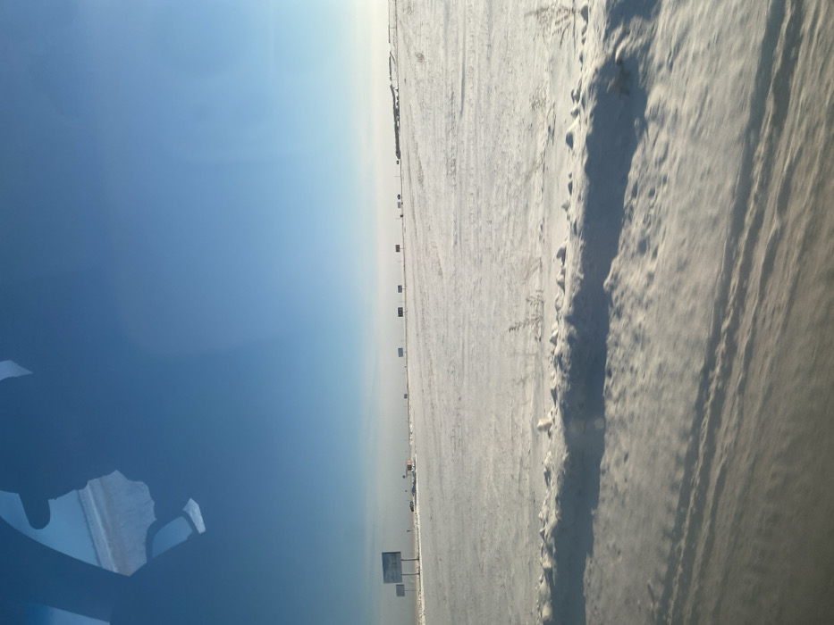 This image depicts a vast, snow-covered landscape identified as being in the vicinity of Ozerne, Russia, under a clear, bright blue sky. The ground is entirely blanketed in white snow, displaying subtle undulations and cast shadows in the foreground. Along the distant horizon, a linear arrangement of approximately twelve rectangular billboards with blank or indistinct faces extends across the frame. Several indistinct vehicles, consistent with large trucks, are faintly visible on a distant road adjacent to the billboards in the mid-ground, appearing to travel from left to right. Sparse, dry vegetation stalks protrude from the snow in the immediate foreground. The upper left quadrant of the image contains reflections, indicative of the photograph having been captured from inside a vehicle, showing partial contours of a window frame or interior structure. No individuals are discernible within the scene.