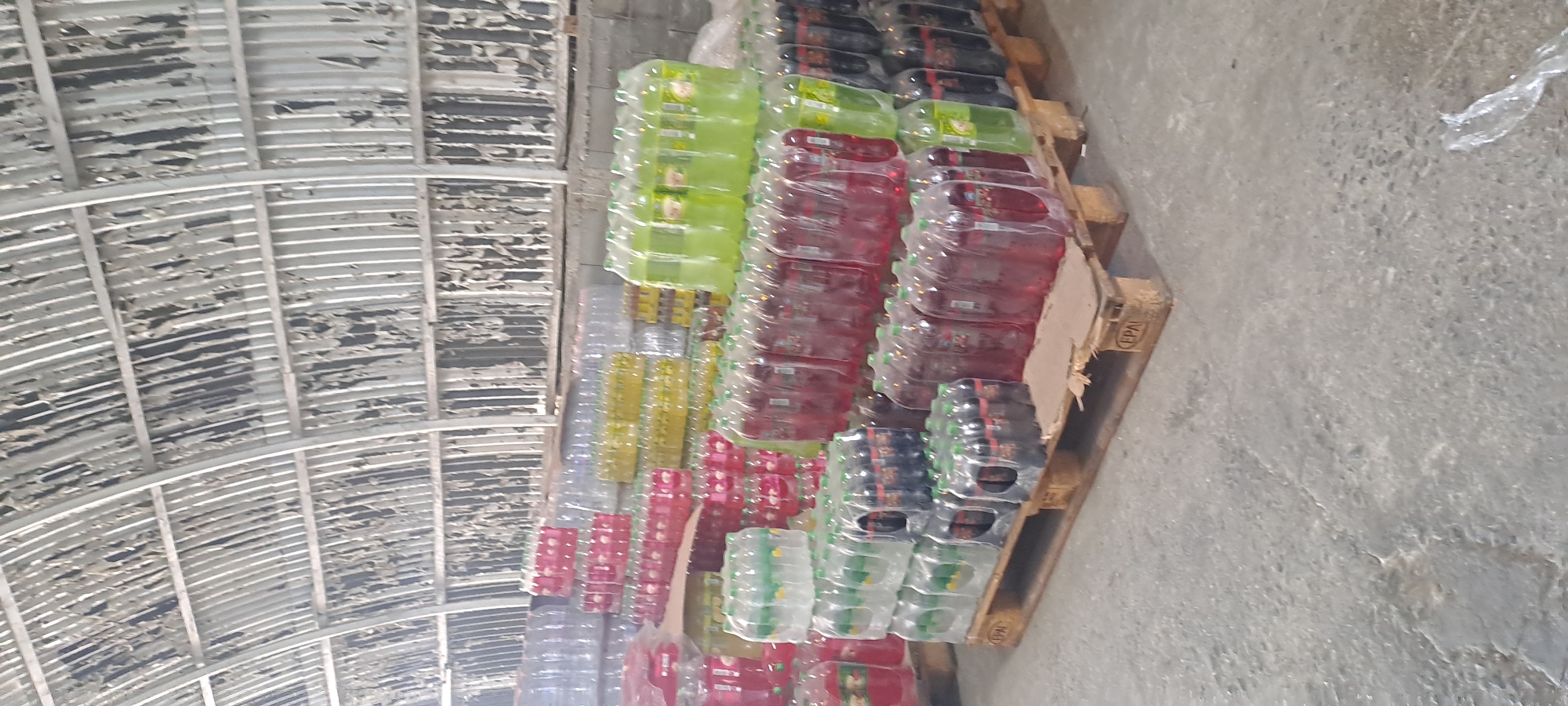 The image shows a warehouse interior in Velika, Bulgaria, with stacks of bottled beverages on wooden pallets. The warehouse has a corrugated metal arched roof with some damaged insulation material. The bottled drinks are packaged in clear plastic shrink wrap. There are several rows of stacked bottles, predominantly in red, clear, yellow, and green colors, indicating different beverage flavors or types. Some pallets have "EPAL" stamps, indicating they are European Pallets. The floor is concrete. No people are visible in the image. The scene appears to be a storage area for a beverage distribution or manufacturing facility.