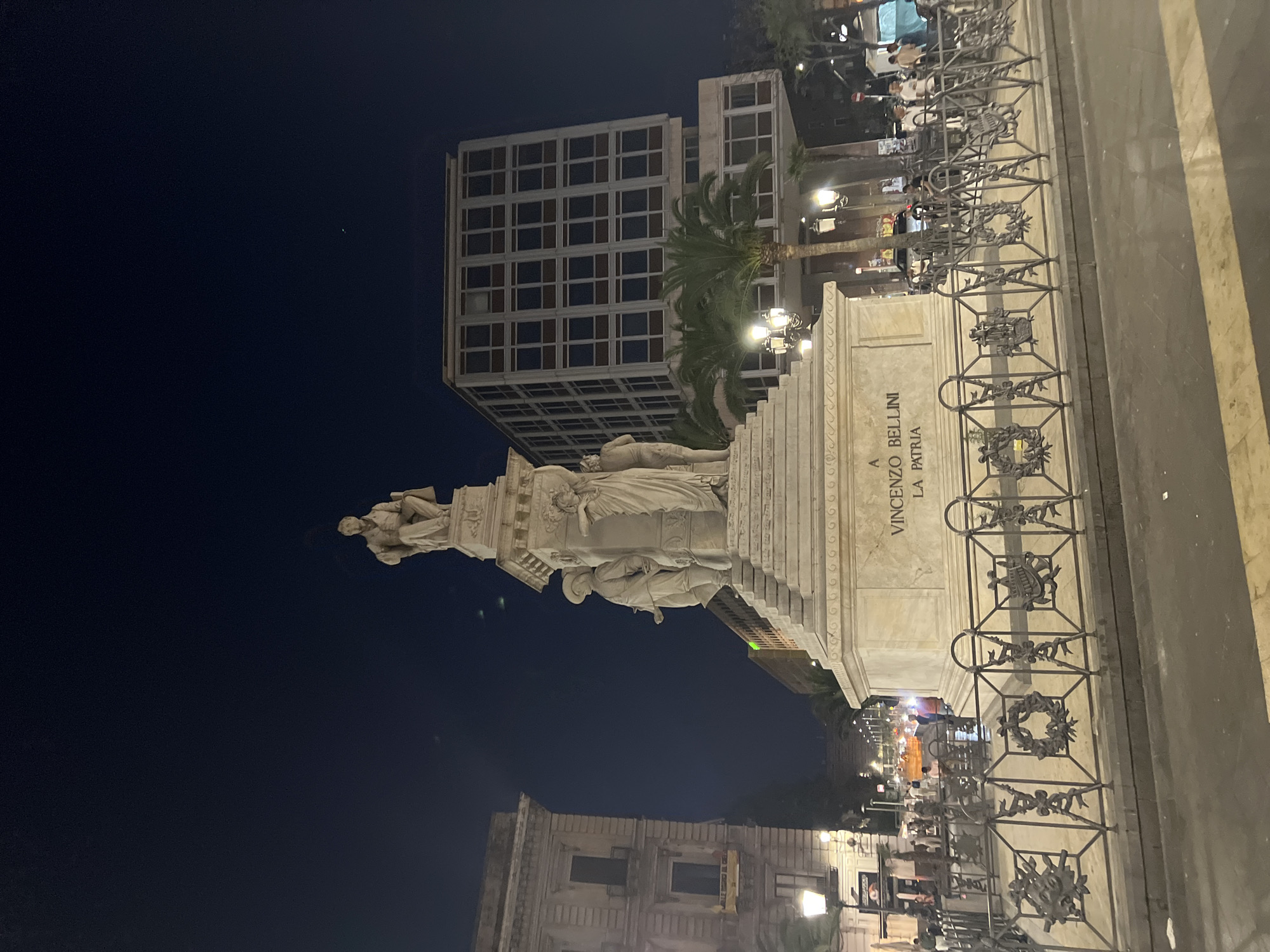 The image shows a monument at night in Catania, Italy, specifically Piazza Vincenzo Bellini, dedicated to Vincenzo Bellini. The monument is a large, white stone structure with multiple statues. The central figure is seated, holding a book. Below this figure are three other statues: two men and a woman. The monument's base is inscribed with "A VINCENZO BELLINI LA PATRIA". The monument is surrounded by a decorative metal fence with various emblems. In the background, there is a modern, multi-story building with many windows, a palm tree, and several street lamps. Several people are visible in the background, some walking and some standing, illuminated by streetlights. The sky is dark, indicating nighttime. The overall scene depicts a public square at night with a prominent monument.