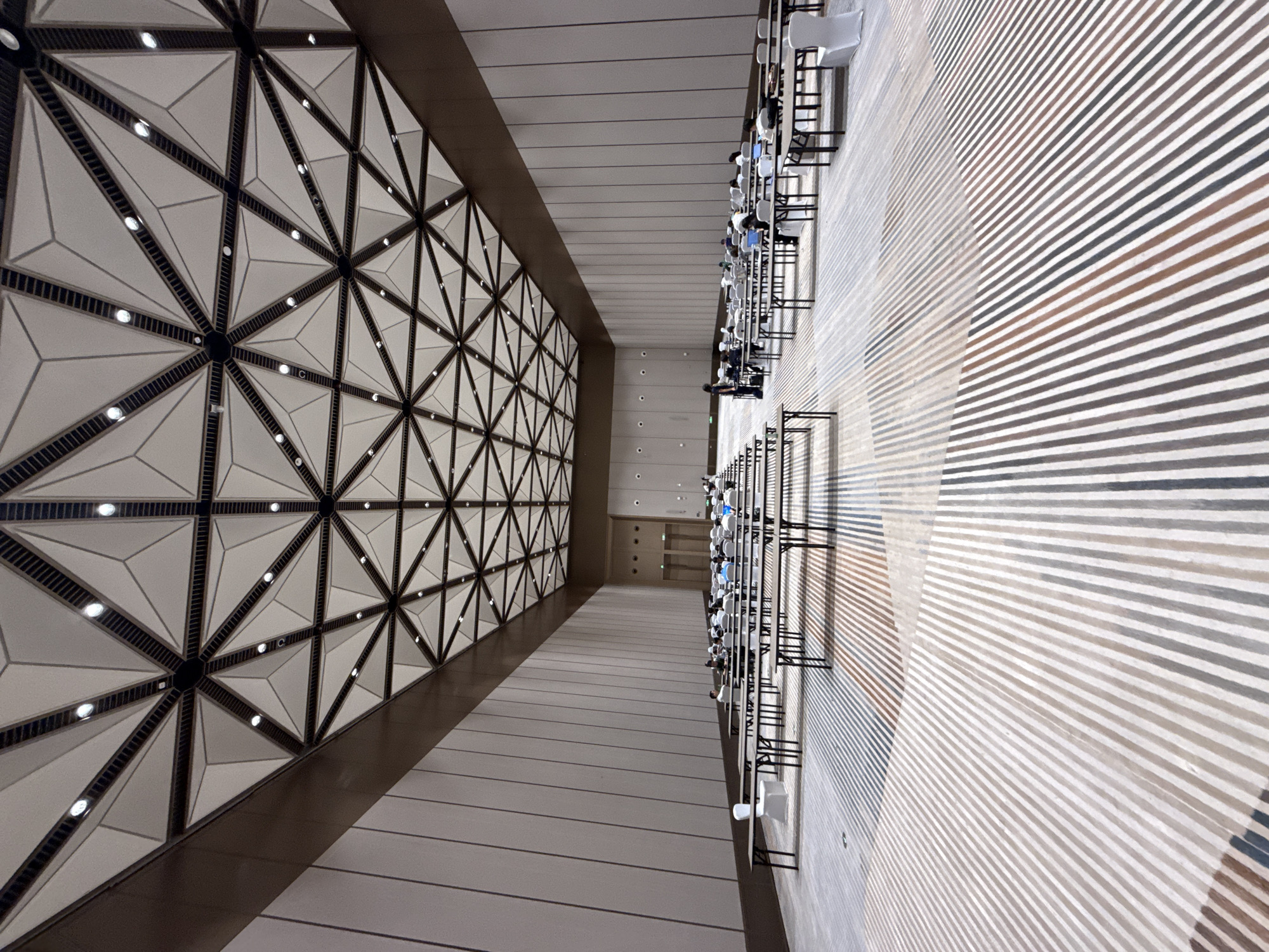 The image shows a large, modern conference hall with a geometric, multi-faceted ceiling and a striped carpeted floor. The hall is set up with rows of tables and chairs, suggesting a lecture, presentation, or examination setting. Several individuals are seated at the tables, some appearing to be working on laptops or taking notes. The overall environment is spacious and brightly lit by overhead lighting integrated into the ceiling design. The scene is located in the city of Chongqing High-tech Zone, China.