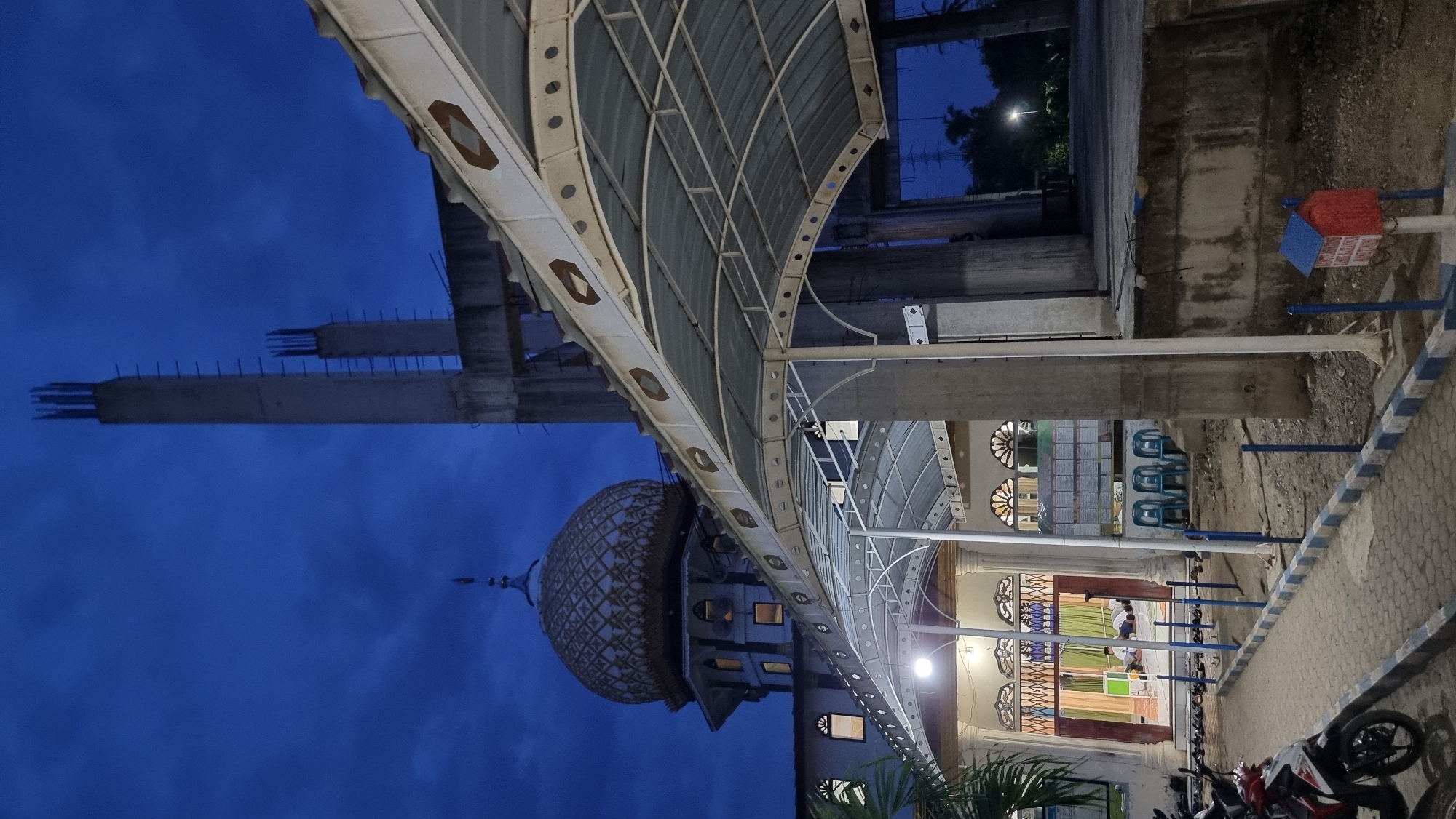 The image depicts the exterior of a mosque under construction or renovation in Manetunong, Indonesia, at dusk. A prominent dome with intricate golden patterns is visible on the mosque's main structure, alongside unfinished concrete pillars and beams suggesting ongoing construction. A covered walkway with a corrugated roof extends towards the viewer, supported by white pillars. In the background, a tall, unfinished concrete tower with exposed rebar rises into the deep blue, cloudy sky.
In the foreground, a paved walkway lined with blue and white borders leads towards the mosque's entrance. Several motorcycles are parked along the left side of the walkway. The entrance to the mosque is open, revealing an interior with several people engaged in prayer, indicated by their kneeling postures. Blue chairs are placed outside the entrance. A small red and blue mailbox-like structure is situated near the walkway on the right. The overall environment suggests a community gathering space in a semi-urban or rural setting, characterized by a blend of traditional architecture and modern construction.