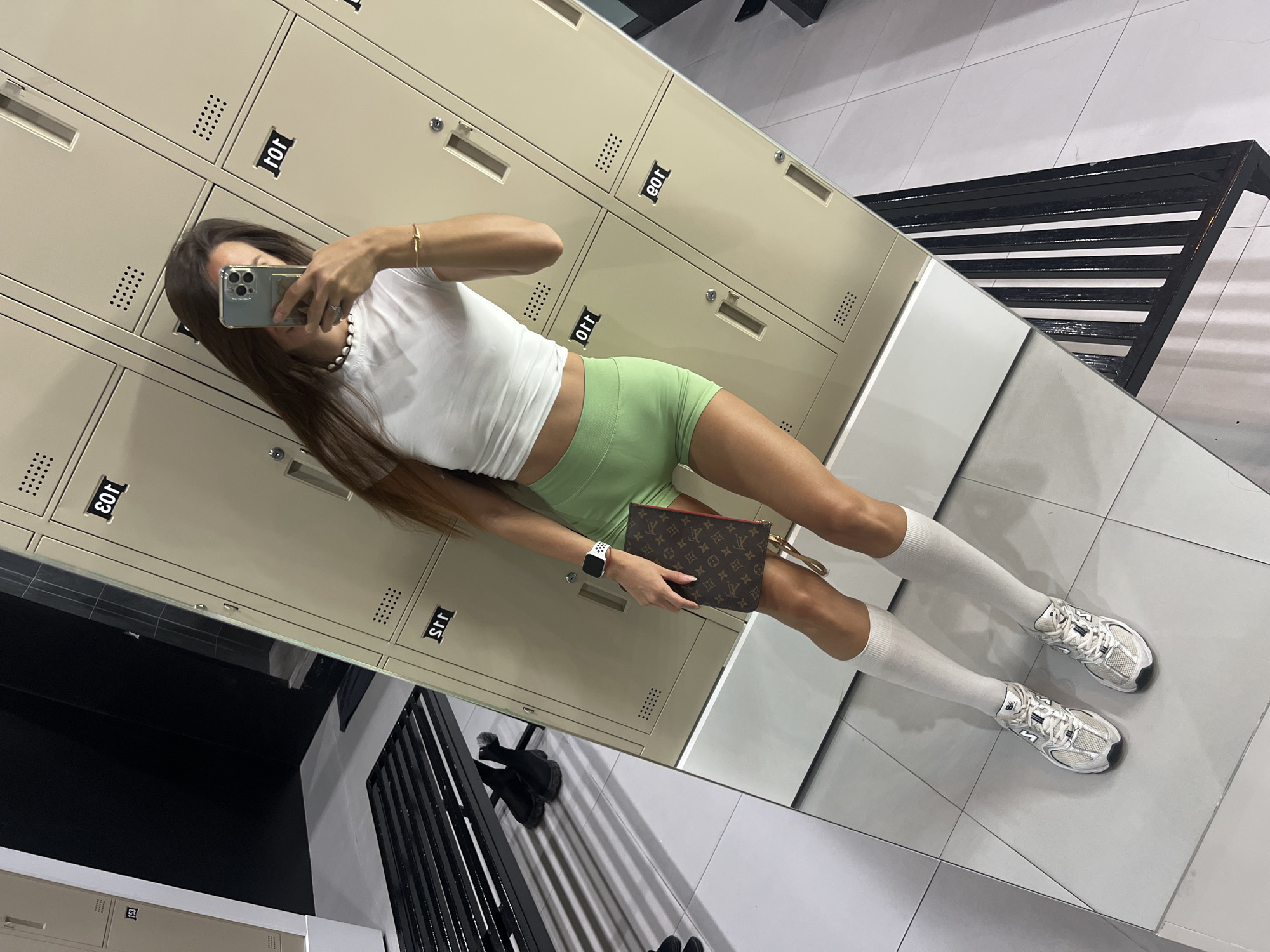 In a locker room, a person is taking a mirror selfie. They are wearing a white crop top, light green shorts, white knee-high socks, and silver athletic shoes. A Louis Vuitton clutch is held in their left hand. The background consists of beige lockers and a black bench. The image is taken from a high angle, with the subject positioned in the center.