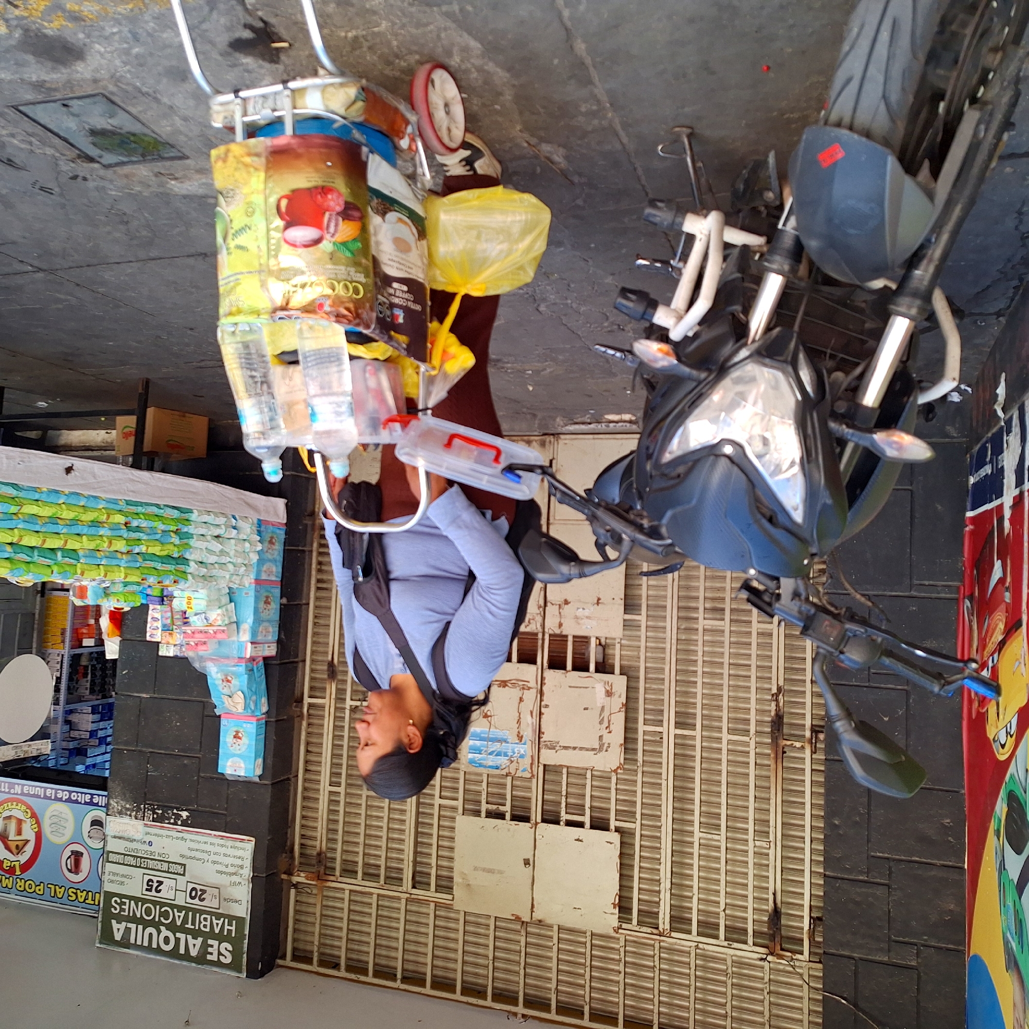 The image depicts a woman standing next to a vending cart and a motorcycle in front of a building with a closed, slatted metal gate. The woman is wearing a light blue shirt and has a black strap across her shoulder. The vending cart is loaded with various items, including bottled water, packages of coffee or cocoa mix, and other goods. Next to the cart is a motorcycle. The storefront next to the gate has advertising signage in Spanish. The location appears to be urban, likely a street in an area where rental rooms are available. The scene is captured in a static, medium shot.