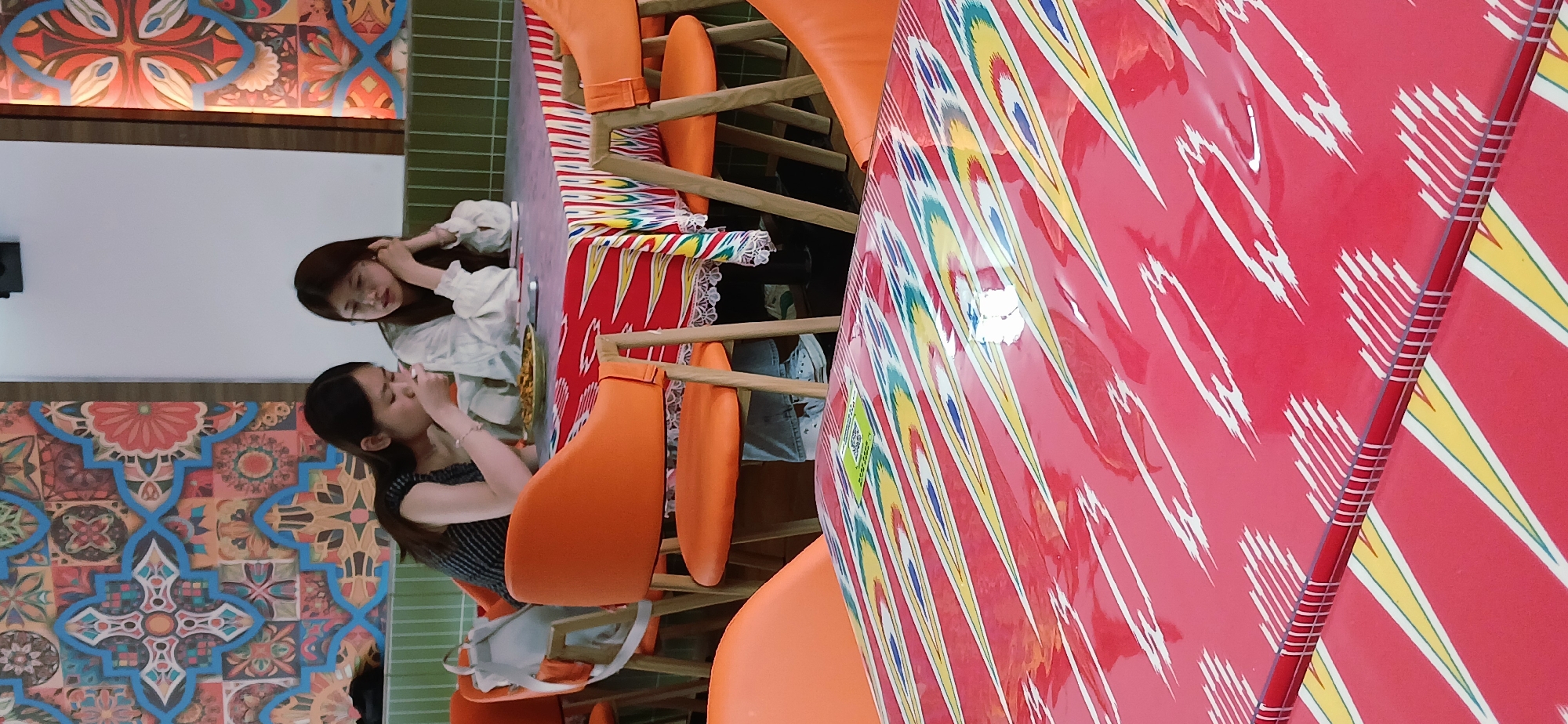 Two young women are seated at a table inside a restaurant. The woman on the left is eating while the woman on the right is adjusting her hair. The table is covered with a colorful cloth. Orange chairs surround the table. The restaurant features colorful tile patterns on the walls. The location appears to be in the Chongqing High-tech Zone, China.