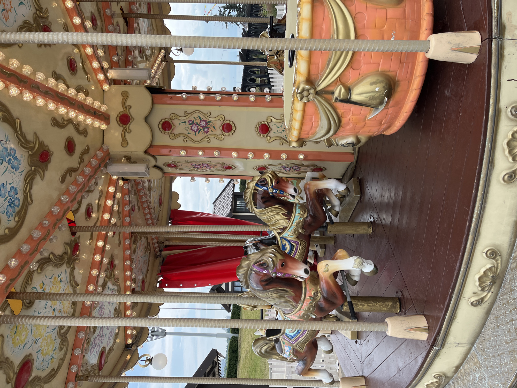 This image shows a close-up view of a carousel horse from a fairground ride, situated outdoors in Nizhny Novgorod, Russia. The horse is painted in shades of brown with a golden mane and tail, and decorated with colorful saddle blankets featuring floral patterns. It is mounted on a brass pole, which is attached to the wooden platform of the carousel. The carousel roof is visible above, adorned with lights and ornate detailing. In the background, glimpses of buildings and greenery suggest an urban park or entertainment venue. The sky is overcast, indicating it is likely daytime.