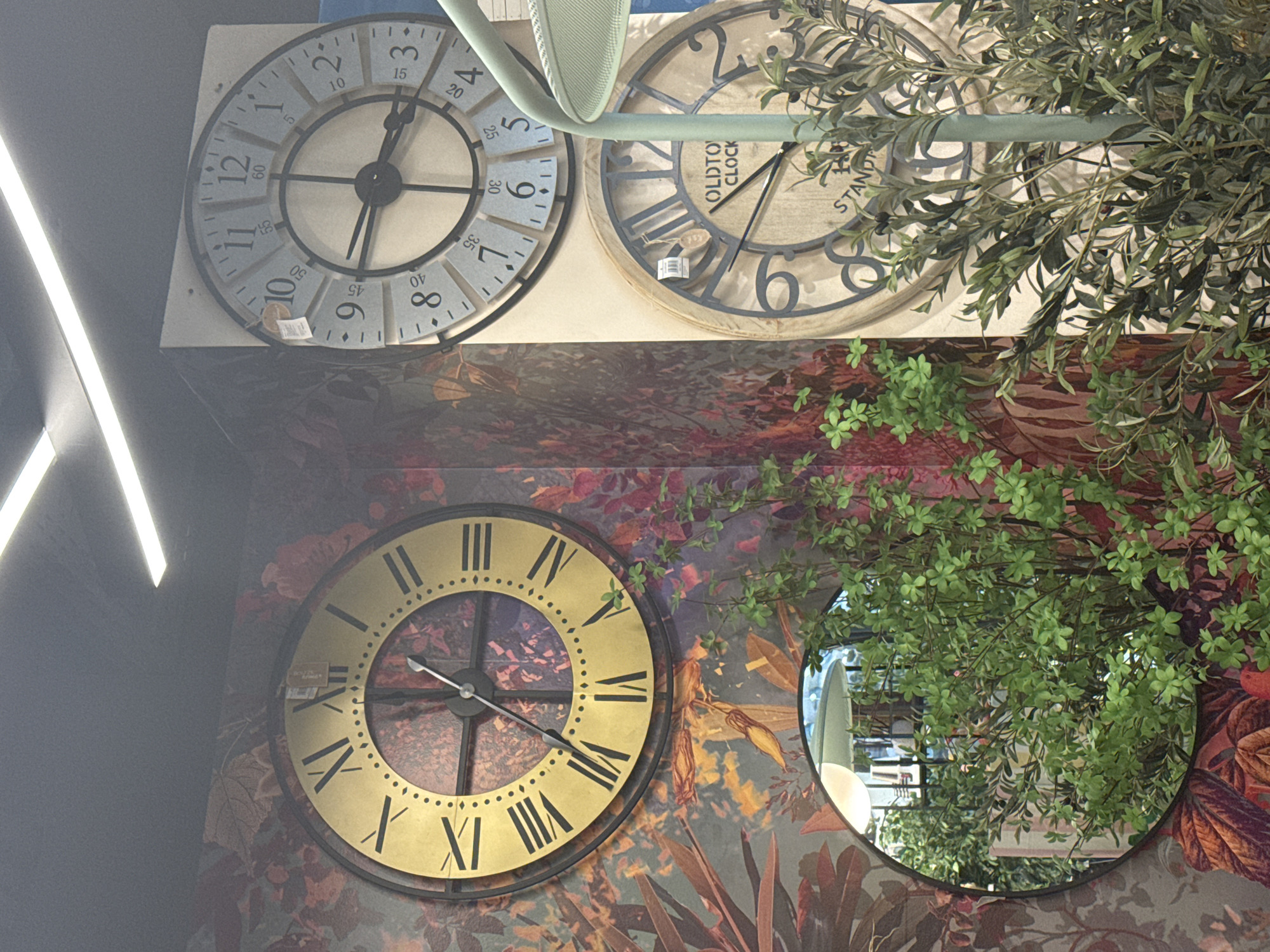 The image shows an interior setting with three large decorative clocks. The clock on the left is gold with black Roman numerals and has a floral patterned background visible through its face. The clock above and to the right is light blue with black Arabic numerals and has minute markers. The clock below and to the right is light brown with black Arabic numerals and appears to be labeled "OLDTOWN CLOCK STANDARD". A circular mirror reflecting greenery and lighting is positioned in the lower left corner. Green plants are visible in front of the clocks and mirror. The overall impression is of a display of home decor items.
