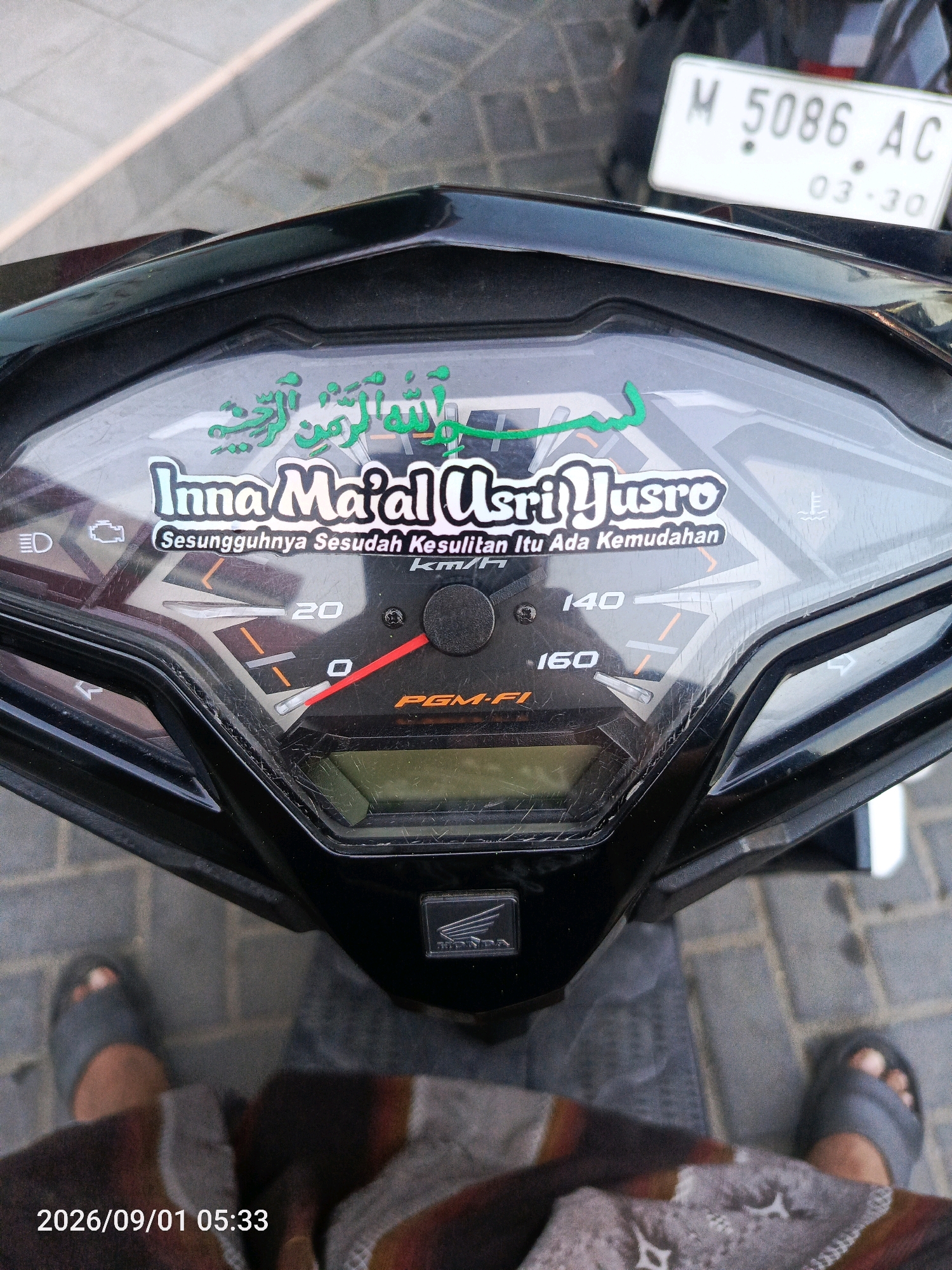 An overhead, eye-level shot depicts the dashboard of a black motorcycle, with a white license plate reading "M 5086 AC 03-30" visible in the background. The motorcycle's dashboard features a speed dial that reads 0-160 km/h, currently indicating a speed near 0 km/h. Green Arabic calligraphy is superimposed on the dashboard, translating to "Inna Ma'al Usri Yusro," meaning "Indeed, with hardship there is ease." Below this, in white text, is "Sesungguhnya Sesudah Kesulitan Itu Ada Kemudahan." The motorcycle is parked outdoors on a paved surface. The timestamp in the lower left corner indicates the image was taken on September 1, 2026, at 5:33 AM. No individuals are clearly visible, but feet wearing gray slippers are present in the lower portion of the frame.