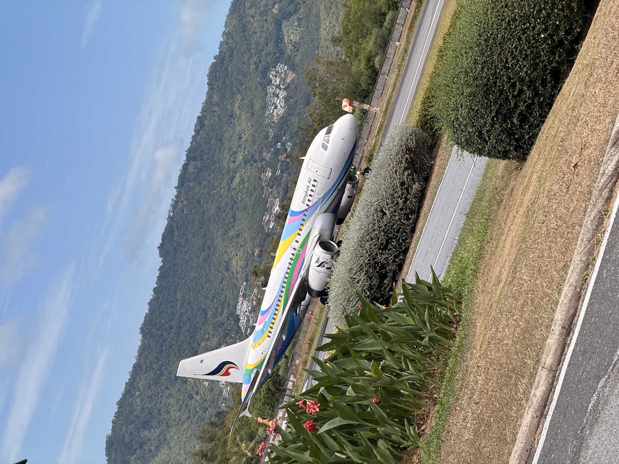 An airplane with a colorful livery, belonging to Bangkok Airways, is on a tarmac, likely during takeoff or landing, at the Samui Airport in Koh Samui, Thailand. The aircraft is positioned at an angle, with its nose pointing slightly towards the viewer. The tail of the plane features the airline's logo and is adorned with vibrant streaks of yellow, pink, blue, and green. The fuselage displays "Bangkok Air" and the registration "HS-PGN".
The foreground of the image is dominated by lush green foliage, with some red flowers blooming amongst the leaves. To the right, a manicured hedge lines the edge of the tarmac. In the background, a lush, green, mountainous landscape rises, dotted with houses. The sky above is blue with scattered, wispy clouds. The overall scene suggests a sunny day in a tropical location. The airport appears to be situated in a natural setting, with the mountains providing a scenic backdrop. The image captures a dynamic moment of aviation against a beautiful natural environment.