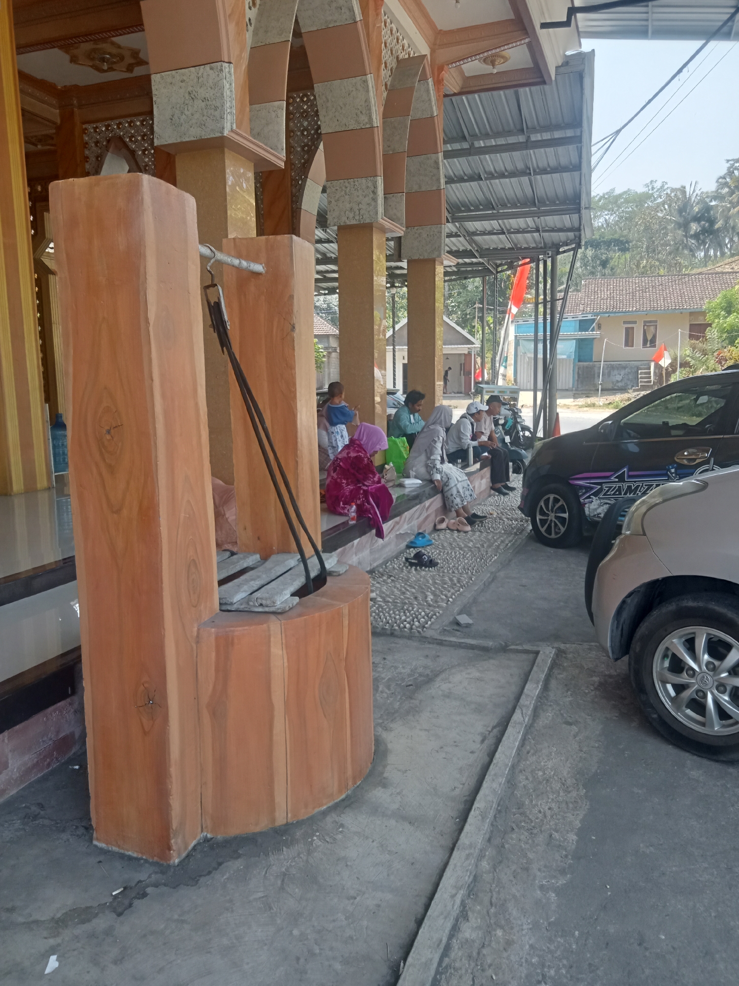 The image captures an outdoor scene in Tlogosari, Indonesia, featuring a water well structure in the foreground constructed from light brown wooden planks. The well has a metal bar across its opening with two black ropes attached, suggesting it is a functional well. Several people are seated on a raised platform to the right of the well, some conversing. Behind them, two cars are parked. One car, a dark SUV with a purple and silver decal, is partially visible. The other car, a lighter colored vehicle, is in the lower right corner. In the background, a building with a tiled roof is visible, along with trees and utility poles. The overall impression is a public space, possibly near a place of worship or community center, with people gathered and vehicles parked. The lighting suggests it is daytime.