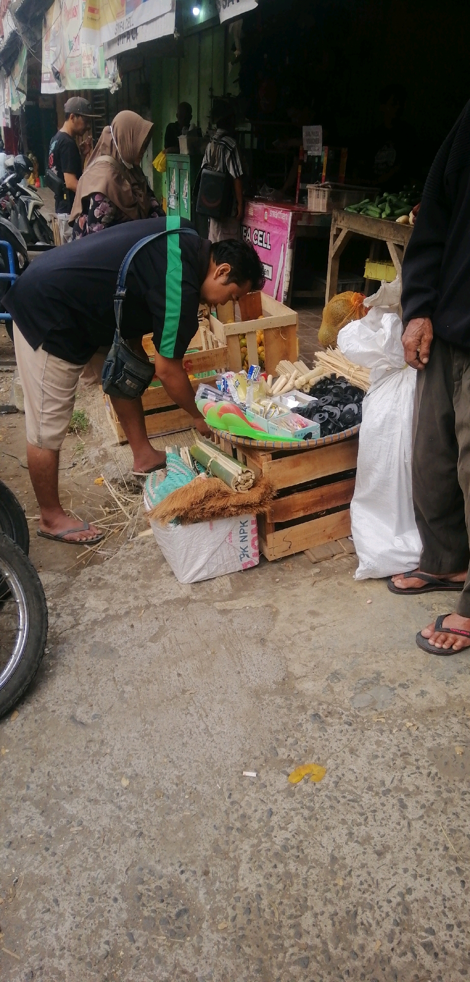 The image depicts a street-level market scene in Ajibarang, Indonesia. In the foreground, a man in a black shirt with green trim and beige shorts is bent over, examining items on a woven tray. He is wearing a crossbody bag. To his left, a motorcycle wheel is partially visible. Behind him, several wooden crates are stacked, some containing produce like oranges, and others holding various goods such as small white tubes, black circular objects, and packaged items. To the right, another man in dark pants and flip-flops stands with his hand on a large white bag. Further back, other individuals, including a woman in a hijab, are present near market stalls. The environment appears to be an outdoor marketplace with a concrete ground surface.