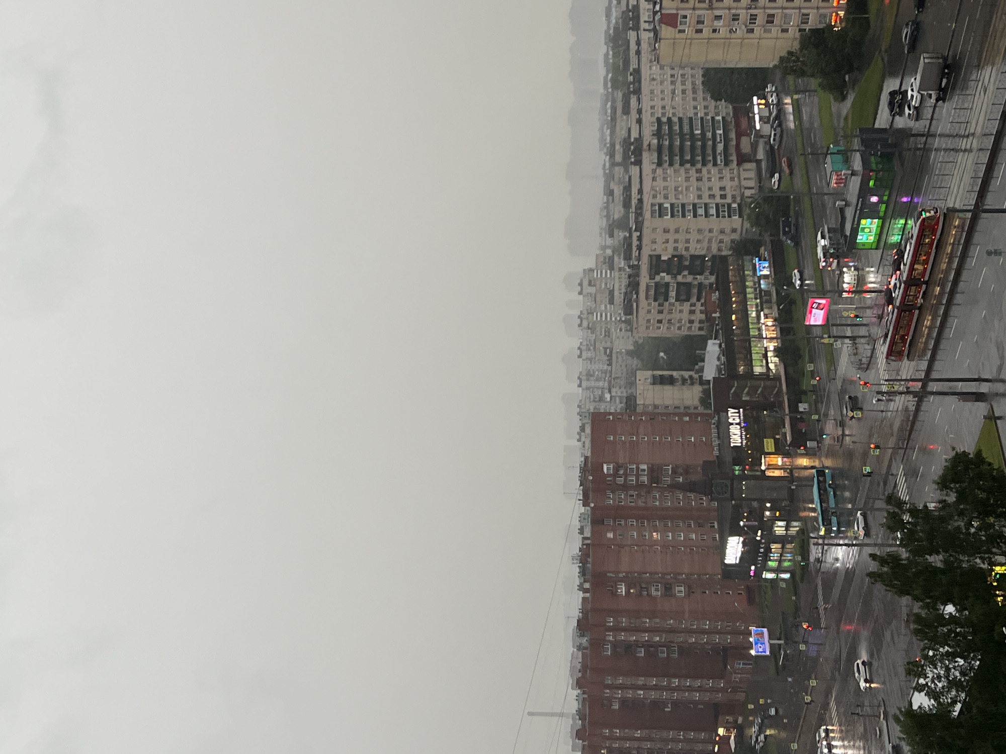 This is a horizontal, eye-level, medium shot of a city street scene in the rain. The sky is overcast and hazy, with a light gray color dominating the upper portion of the frame. In the foreground, a wet asphalt street reflects the lights of buildings and vehicles. Several cars and a truck are visible on the street, as well as a red tram with its lights on. Traffic lights are red and green. Buildings of various heights and architectural styles line the street. Some buildings have illuminated windows and signs. A large red brick building occupies the left side of the frame, and in the center, a building with a dark spire is visible. The overall impression is one of a damp, urban environment.
