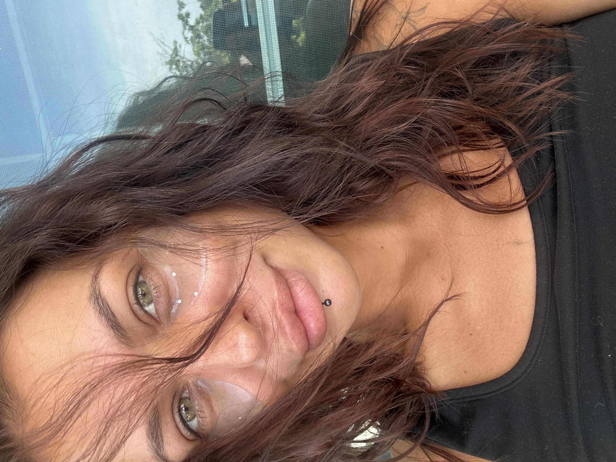 A close-up, eye-level, medium shot captures a woman's face and upper chest, viewed from the left shoulder. Her long, dark brown hair is slightly disheveled, framing her face. She is wearing a black tank top. Under her eyes are clear eye patches with small, silver star-shaped glitter accents. A small, black, circular stud is visible in her lower lip. The background is out of focus, showing a blue-tinted window with a mesh screen and some greenery beyond, suggesting an outdoor or semi-outdoor setting. The lighting is natural, casting subtle shadows. The overall impression is casual and unposed. The image was likely taken in Ko Samui District, Thailand, given the context.