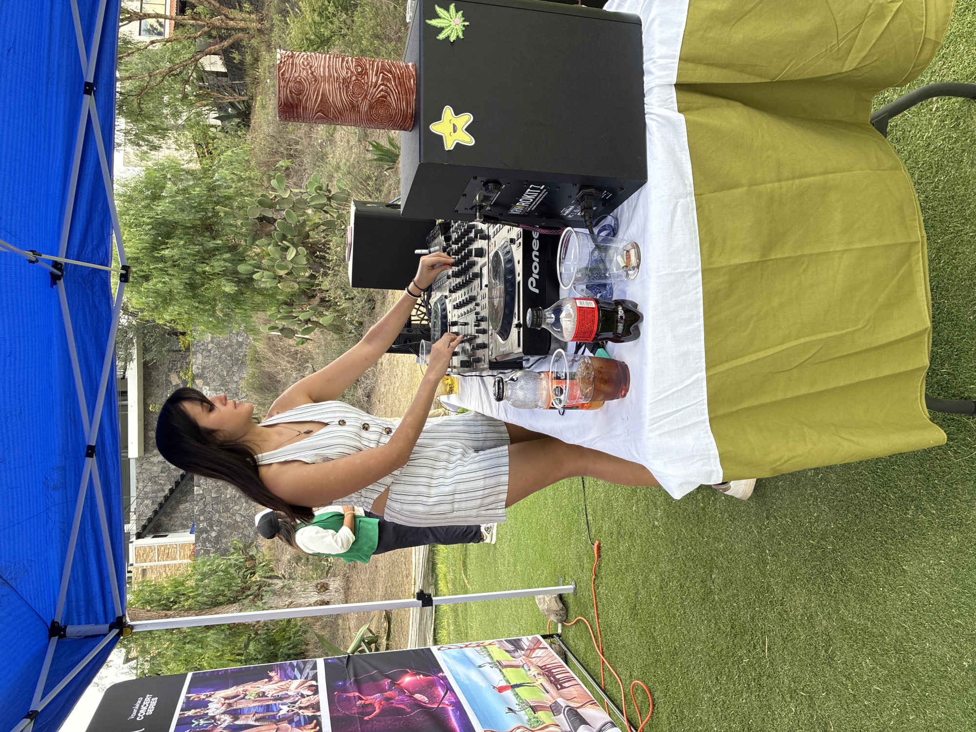 A young woman is operating a Pioneer DJ controller set up on a table with a white and olive green tablecloth outdoors in Pueblo Nuevo de Jasso, Mexico. The DJ equipment is positioned in front of a black speaker with a marijuana leaf and a star sticker on it, as well as a wood grain cylinder. Clear plastic cups and bottles of Coca-Cola and an unknown beverage are also on the table. The DJ is wearing a white and light blue striped dress. A green vest is visible on a person behind her. A blue tent frame stands to the left with promotional concert posters displayed below. The location appears to be a grassy area, with a landscape containing trees, shrubbery, and buildings in the background.