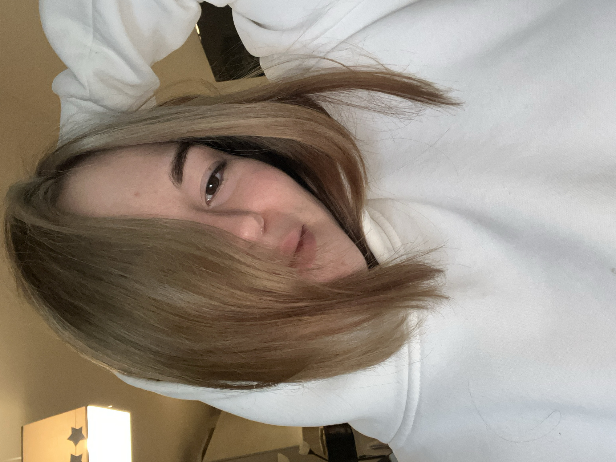 The image displays a close-up, slightly high-angle portrait of a single female individual indoors in Boreti, Montenegro. The subject possesses light brown, shoulder-length hair, styled to partially obscure her left eye and nose. Her visible right eye is dark brown and directed towards the camera. Her lips are slightly parted. The individual is wearing a white hooded sweatshirt, the hood of which is pulled over her head and appears to be held in place by both hands, with the left hand visible near the top of the hood. In the upper-left quadrant of the background, an illuminated square-shaped ceiling lamp with a light-colored shade is visible, featuring two distinct dark star-shaped cutouts. The immediate background is blurred, consisting of warm-toned walls or ceiling in the upper portions and indistinct darker elements below, suggesting an interior room illuminated by artificial light.
