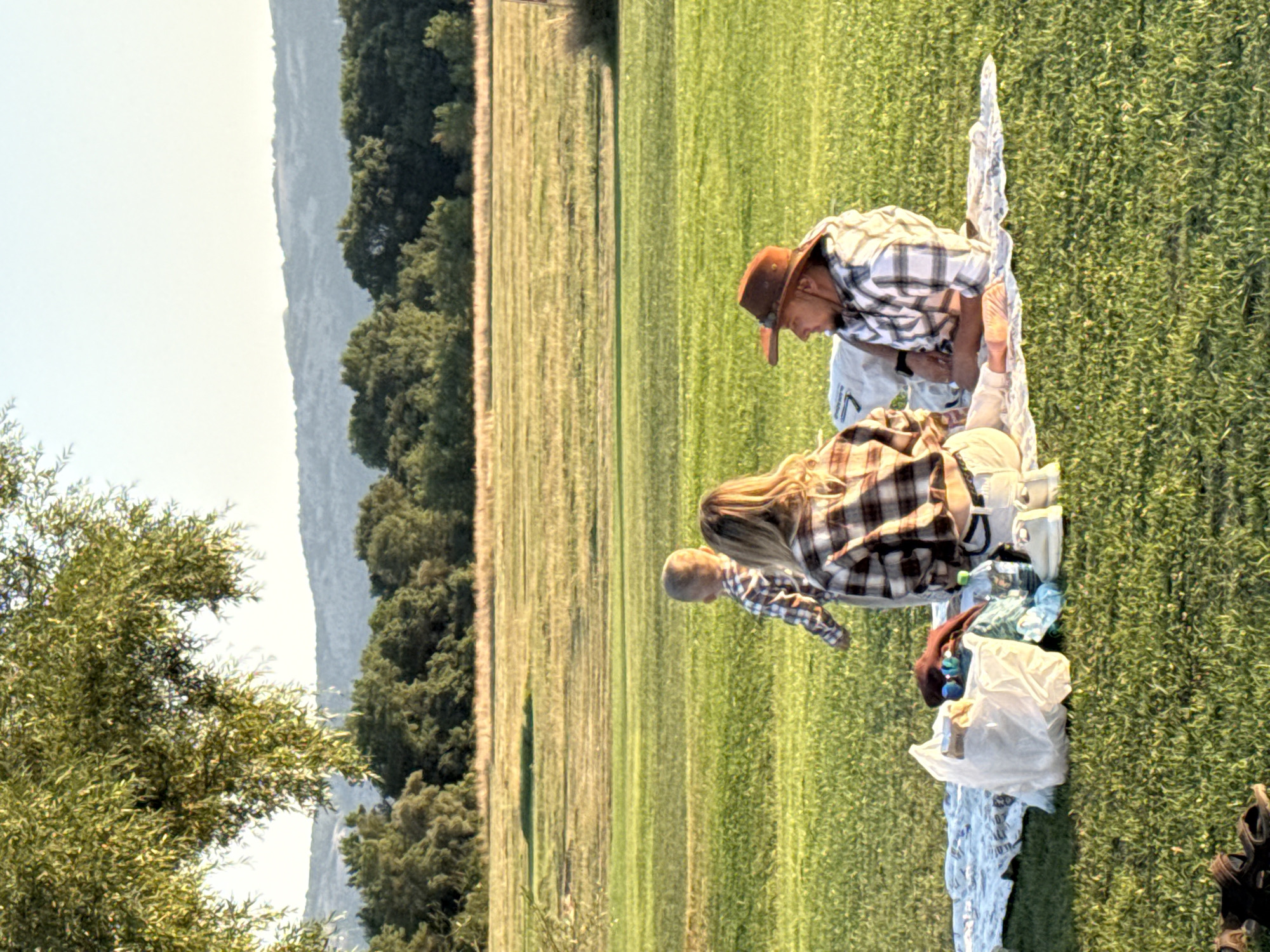 A family is having a picnic in a grassy field. A woman and a man are sitting on a blanket, facing each other. A young child is standing behind the woman, facing away from the camera. There are several items on the blanket, including a white plastic bag, a brown bag, and a water bottle. In the background, there is a field of tall grass, followed by a line of trees and hills. The sky is clear and the sun is setting. The location is identified as Orlynivskyi District, Ukraine.