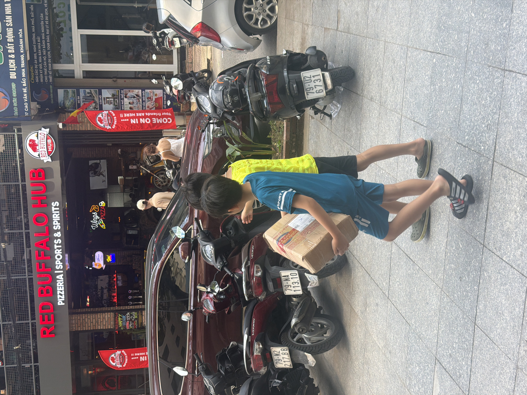 This image captures a vibrant daytime street scene in Bac Nha Trang Ward, Vietnam. The setting is a paved sidewalk alongside a street, lined with buildings housing businesses.
In the foreground, two young boys are walking. One boy, wearing a blue t-shirt and shorts, is carrying a cardboard box labeled "HÀN QUỐC" (meaning South Korea) in his left arm. The other boy, partially visible and wearing a yellow t-shirt, walks slightly ahead of him. Both are wearing sandals.
The street is busy with parked vehicles. Several motorbikes/scooters are visible, including a dark red Honda Vision with license plates "79-NA 113.10" and "79-N2 217.88," and a black scooter with license plate "79-02 6731." A white car is also parked further down the street. In the middle ground, a woman in a white top is bending over a parked scooter.
The background features several storefronts. Prominently displayed is "RED BUFFALO HUB PIZZERIA | SPORTS & SPIRITS" on a large sign, with "RED BUFFALO HUB" logos also on flags and banners. One flag reads "COME ON IN .... Your Friends ARE HERE!!". Other visible signs include "Wings PIZZA," "RED BUFFALO HAPPY HOUR," and "Yes!". Above Red Buffalo Hub is a larger sign for "CAO SƠN GROUP DU LỊCH & BẤT ĐỘNG SẢN NHA T," listing services in Vietnamese such as "BÁN CÁC DỰ ÁN BĐS, NGHỈ DƯỠNG, NHÀ PHỐ, ĐẤT NỀN," and "NHẬN KÝ GỬI, MUA BÁN NHÀ ĐẤT." The general atmosphere suggests a typical bustling urban area.