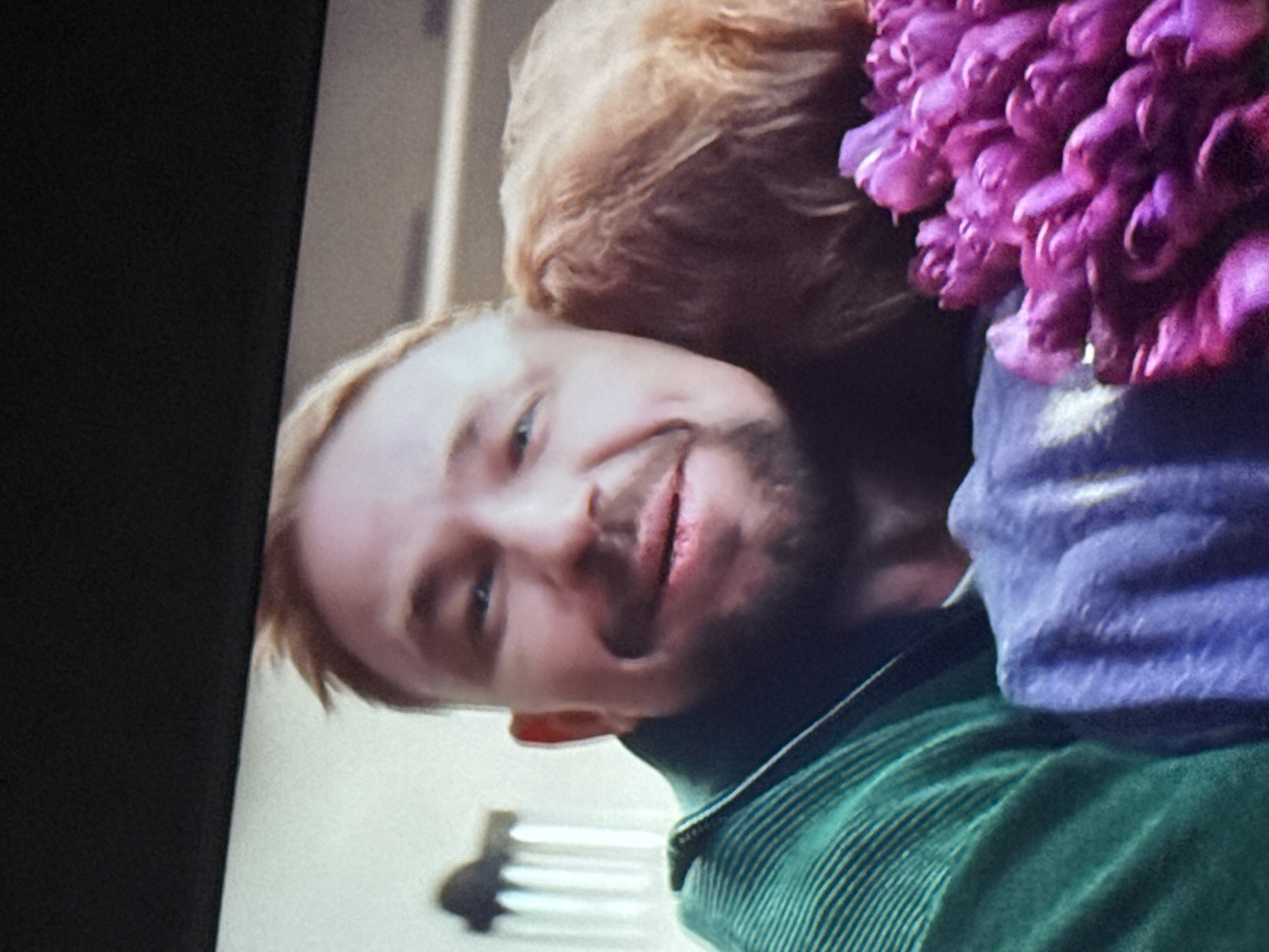 A medium shot shows a man with light brown hair and a trimmed beard smiling slightly. He is wearing a green striped shirt with a dark collar. To the right of his head is a person with curly, light brown hair wearing a light blue garment with a large bouquet of purple flowers. The background is a light, neutral color with a dark, out-of-focus object. The image is taken from a television or monitor. There is no information to suggest this image was taken in Mytishchi, Russia.