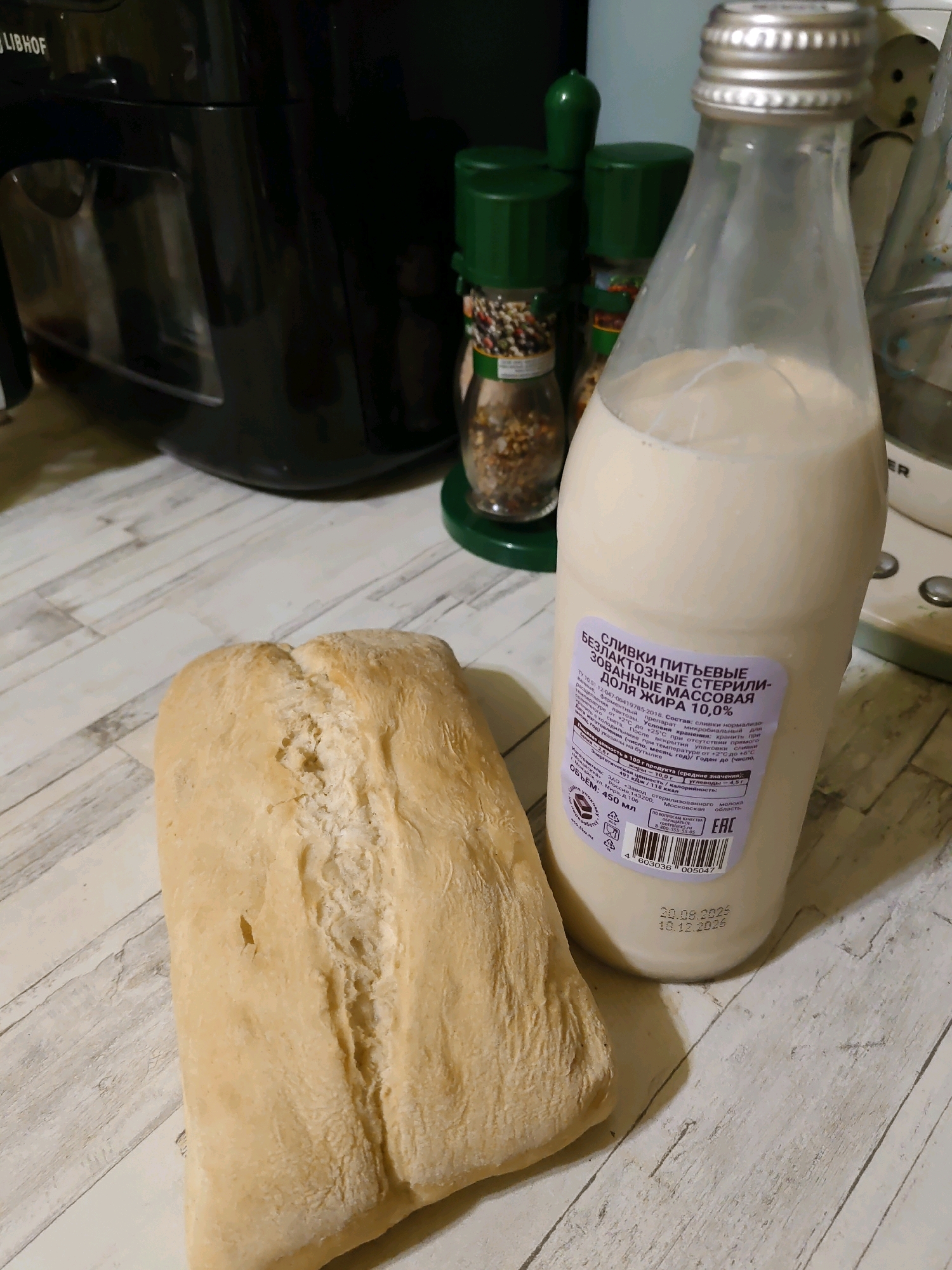 The image displays a close-up, eye-level view of a loaf of bread and a bottle of cream on a wooden table. The bread, golden brown and crusty, is positioned in the foreground, slightly to the left. To its right stands a clear glass bottle with a silver cap, containing a pale, creamy liquid. A label on the bottle has Cyrillic text and a barcode. In the background, out of focus, are kitchen appliances and spice grinders. The lighting is ambient, suggesting an indoor setting. The scene is static and appears to be a still life composition. No specific location within Rostov-on-Don, Russia, can be determined from the provided image.