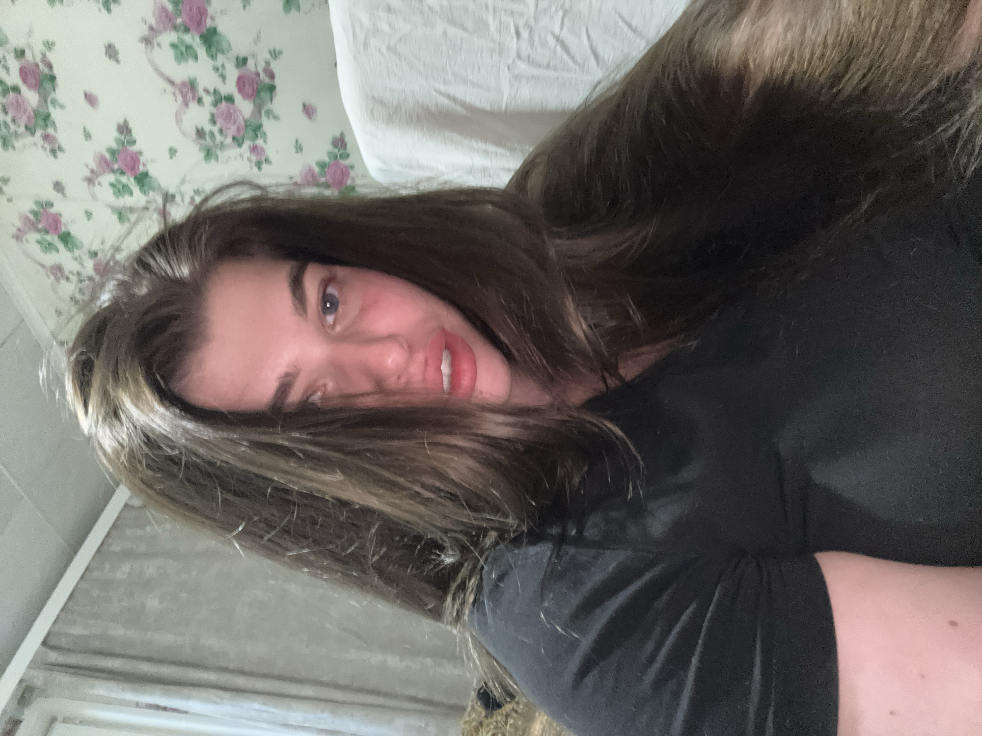 A woman with long, dark brown hair is lying down, facing the camera. She is wearing a black t-shirt. Behind her are a white pillow and a wall with floral wallpaper. The location of the photo is unknown.