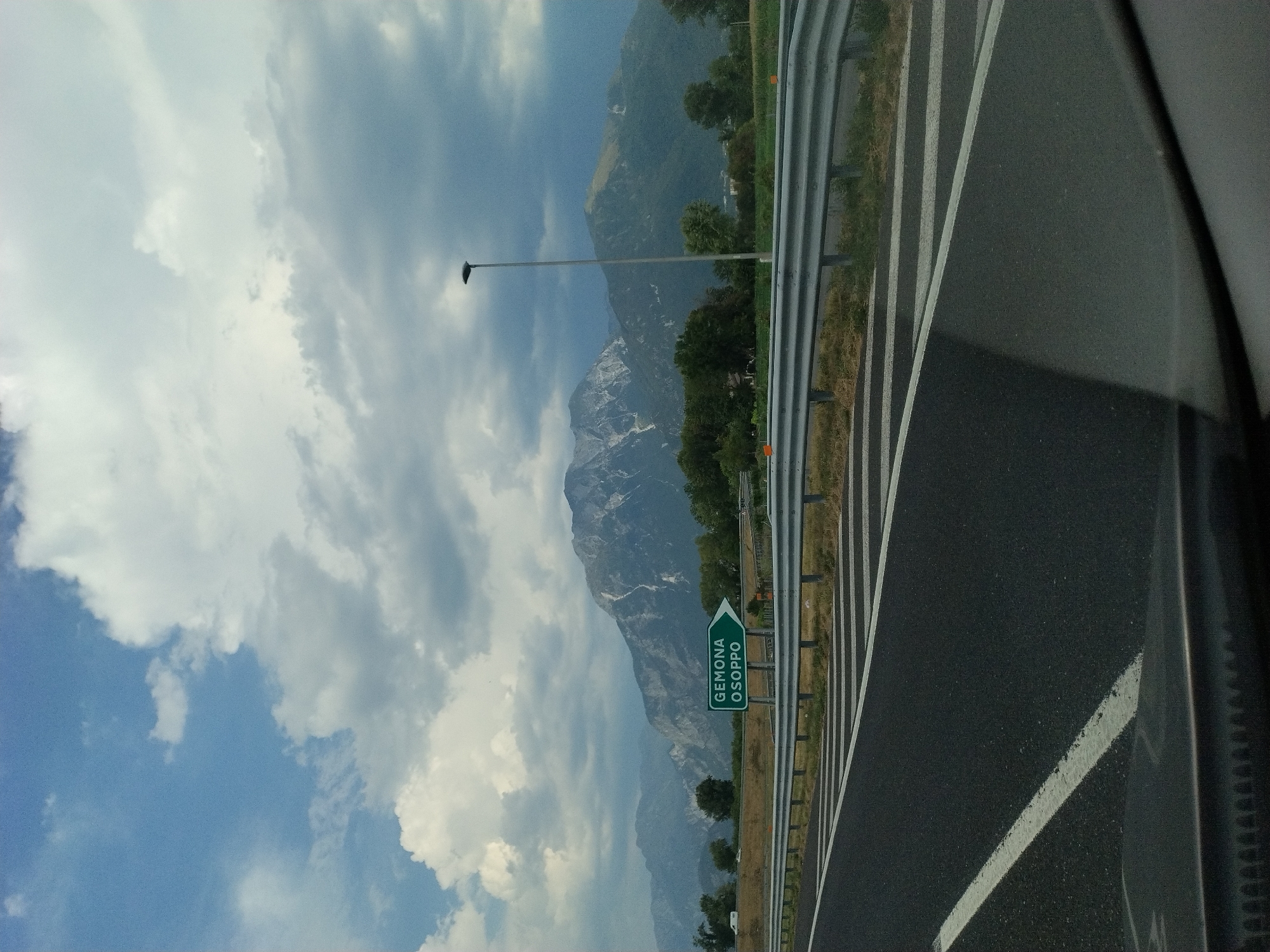 A green directional sign for "GEMONA OSOPPO" points to the right, indicating the direction of travel. The sign is positioned on the side of a multi-lane highway with a metal guardrail separating the traffic lanes from the shoulder. The road surface is asphalt with white lane markings. In the background, a range of rugged, rocky mountains with some patches of green vegetation rises against a cloudy sky. A tall street lamp stands to the right of the sign. The overall scene suggests a view from a vehicle moving along a highway, with a focus on road signage and the mountainous landscape.