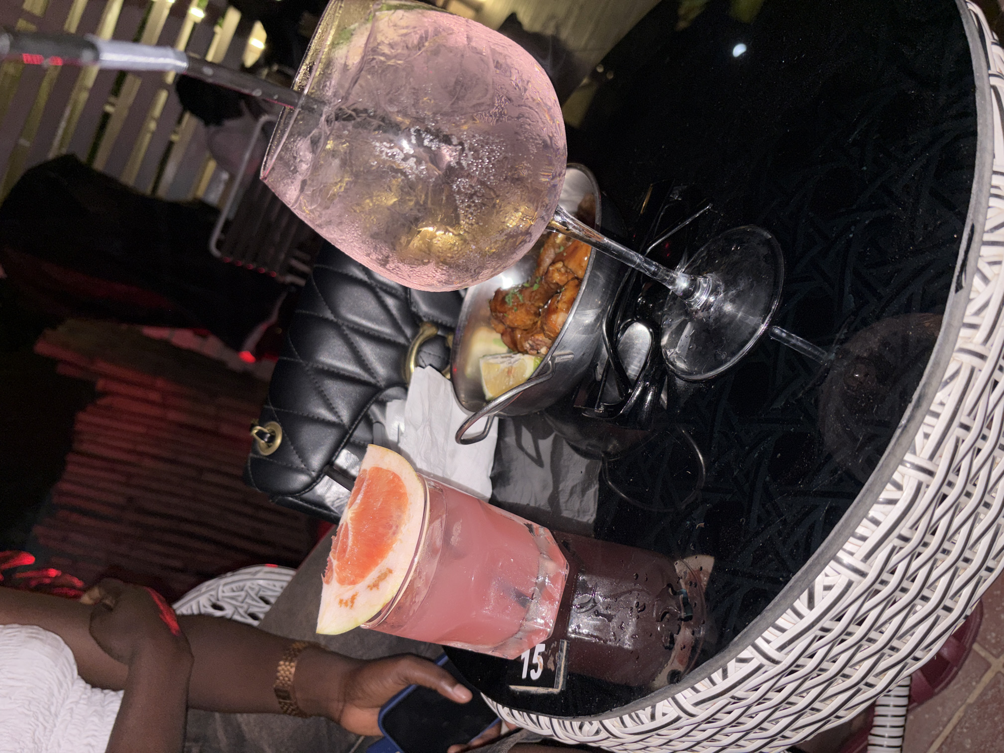 The image shows a table at an outdoor venue in Abuja, Nigeria. A person is seated at the table, holding a smartphone. On the table are two cocktails, a metal bowl containing food (possibly chicken), a black quilted handbag, cutlery, and a table number '15'. The table has a woven wicker base and a black reflective top. The background includes a white picket fence.