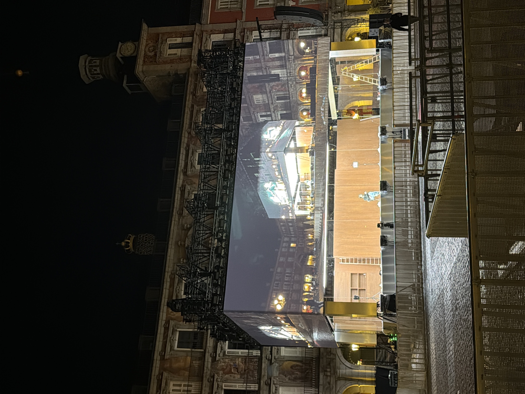 The image shows a nighttime scene in Madrid, Spain, specifically the Plaza Mayor. The photo is taken at an angle, showing the illuminated facades of the buildings that surround the square. There is scaffolding erected, along with a large screen displaying an image of a similar-looking building at night. The scaffolding partially obscures the original building facade behind it. Several metal structures including a grated one are present. A few people are visible on the structures. The background sky is dark, with a single lit structure visible on the top left. The overall lighting is provided by artificial light sources.