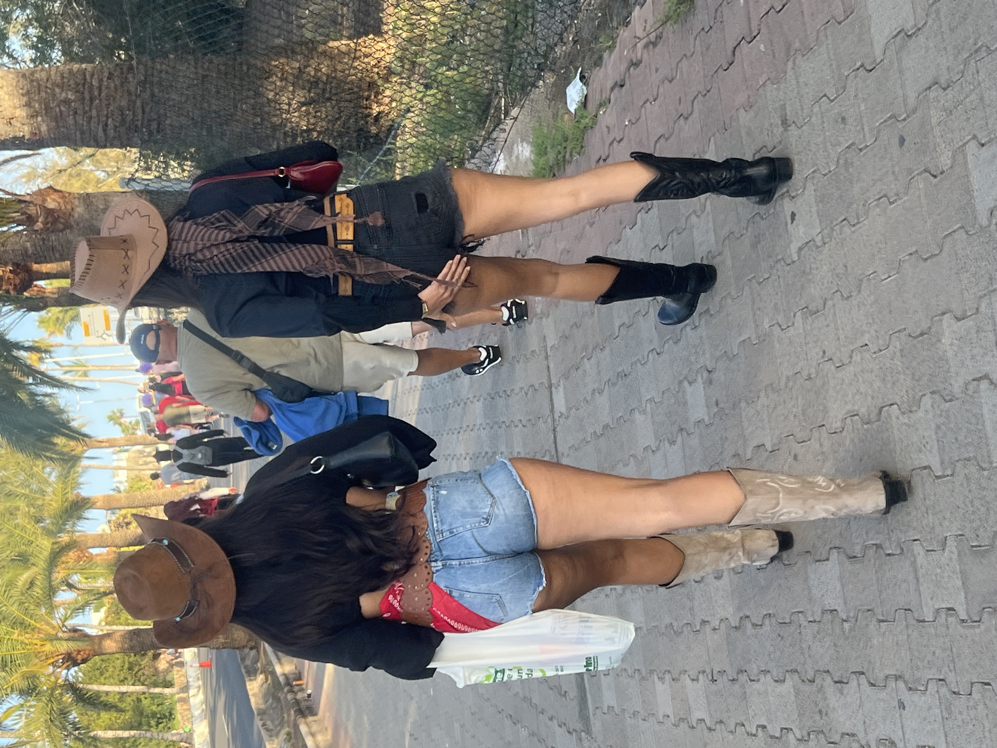 Two women are standing on a sidewalk in San Bartolomé de Tirajana, Spain. Both women are wearing cowboy boots, hats, and denim shorts. The woman on the left is wearing beige boots, a brown hat, and denim shorts with a red bandana. The woman on the right is wearing black boots, a light beige hat, and dark denim shorts with a brown scarf. A group of people walk along the sidewalk in the background. Palm trees are visible in the background. A fence is in the upper left of the frame. The sidewalk is paved with gray stone.