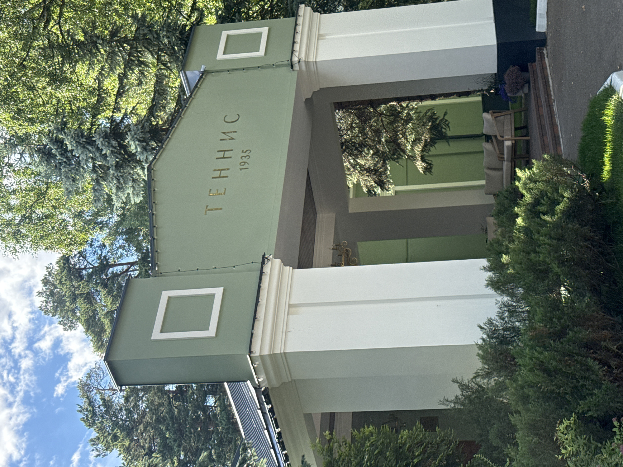 The image shows the entrance to a tennis club in Zhukovka, Russia. The building is painted a light green and white, with the word "ТЕННИС" (Tennis) and the year "1935" inscribed in gold lettering above the entrance. There are two large white pillars on either side of the arched entrance. Inside the entrance, there are two armchairs and a sofa, suggesting a waiting or lounge area. Lush green trees and bushes surround the building, and the sky is blue with scattered white clouds. The scene appears to be set in a well-maintained outdoor environment, likely a park or recreational area.