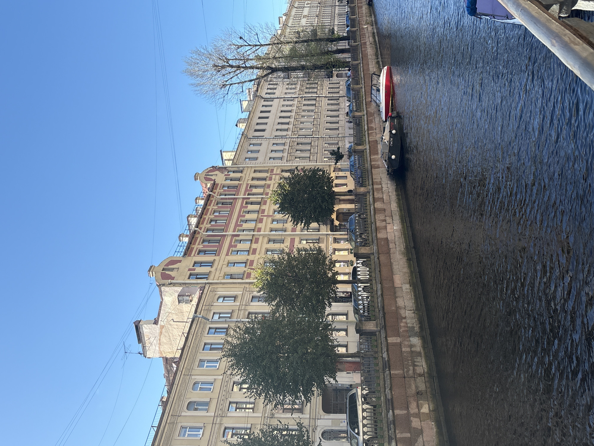 The image depicts a view of a canal lined with historic buildings in Saint Petersburg, Russia. The architecture of the buildings is classical, with multiple stories, arched windows, and decorative facades. Trees are planted along the canal bank, and a metal fence separates the buildings from the walkway. Two boats are moored in the canal, one black with Cyrillic writing on its side and a red and white speedboat. Cars are parked along the street behind the fence. The sky is clear and blue. The scene appears to be a sunny day, likely in the spring or summer based on the foliage of the trees.