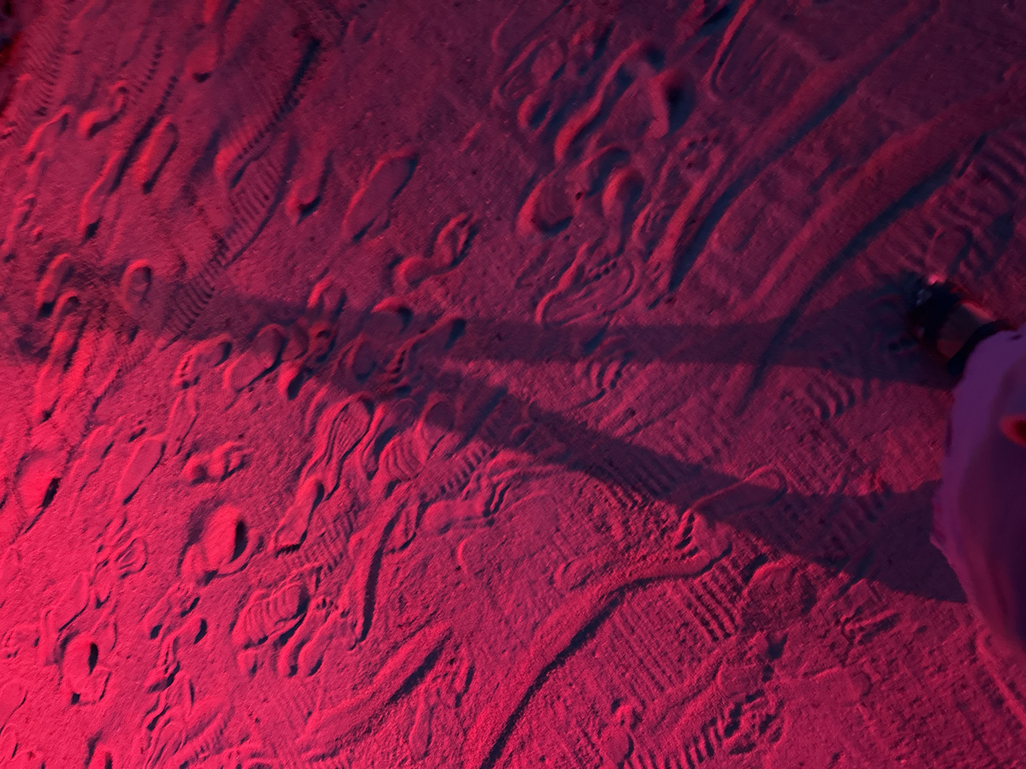 The image shows a close-up, low-angle view of a sandy surface illuminated by a bright pink light. The sand is textured with numerous footprints, some appearing as distinct imprints and others as blurred trails. There is a long, dark shadow cast across the sand, suggesting a person standing in the light source's position. The shadow appears to be that of a human figure, with discernible leg and foot shapes. In the lower right corner of the frame, a dark object, possibly a shoe or bag, is visible at the edge of the sand. The overall scene suggests an outdoor environment, potentially a beach or sandy pathway in Ko Pha-ngan District, Thailand, during nighttime or under artificial lighting.