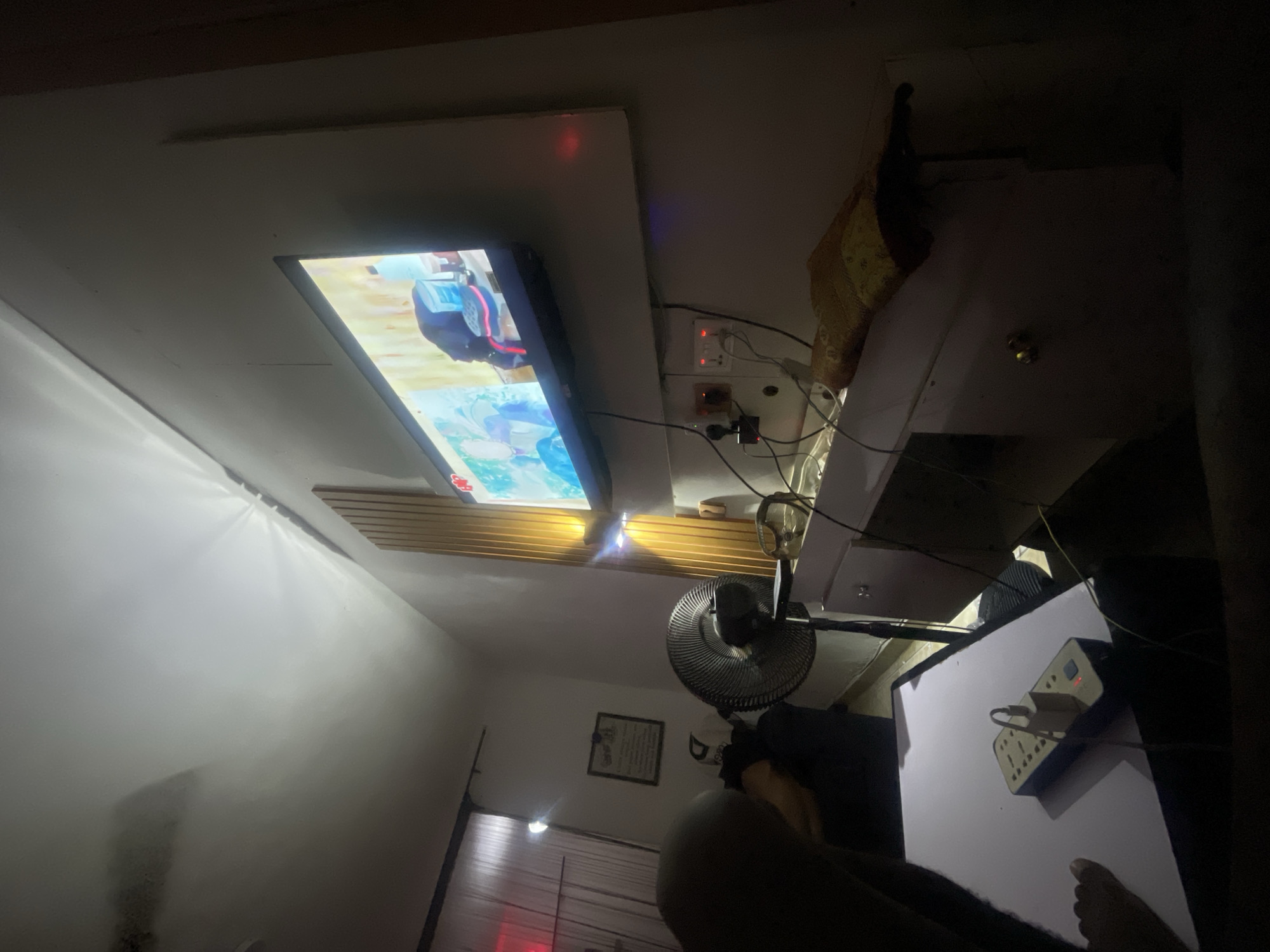 A dimly lit room in a location possibly in Lokoja, Nigeria, contains a flat-screen television mounted on a white surface, displaying an animated image. A standing fan is positioned below the television. Next to the television is a white surface with an electrical outlet strip plugged in. A person is partially visible in the foreground. A window with blinds is visible in the background. A framed picture hangs on the wall near the window.