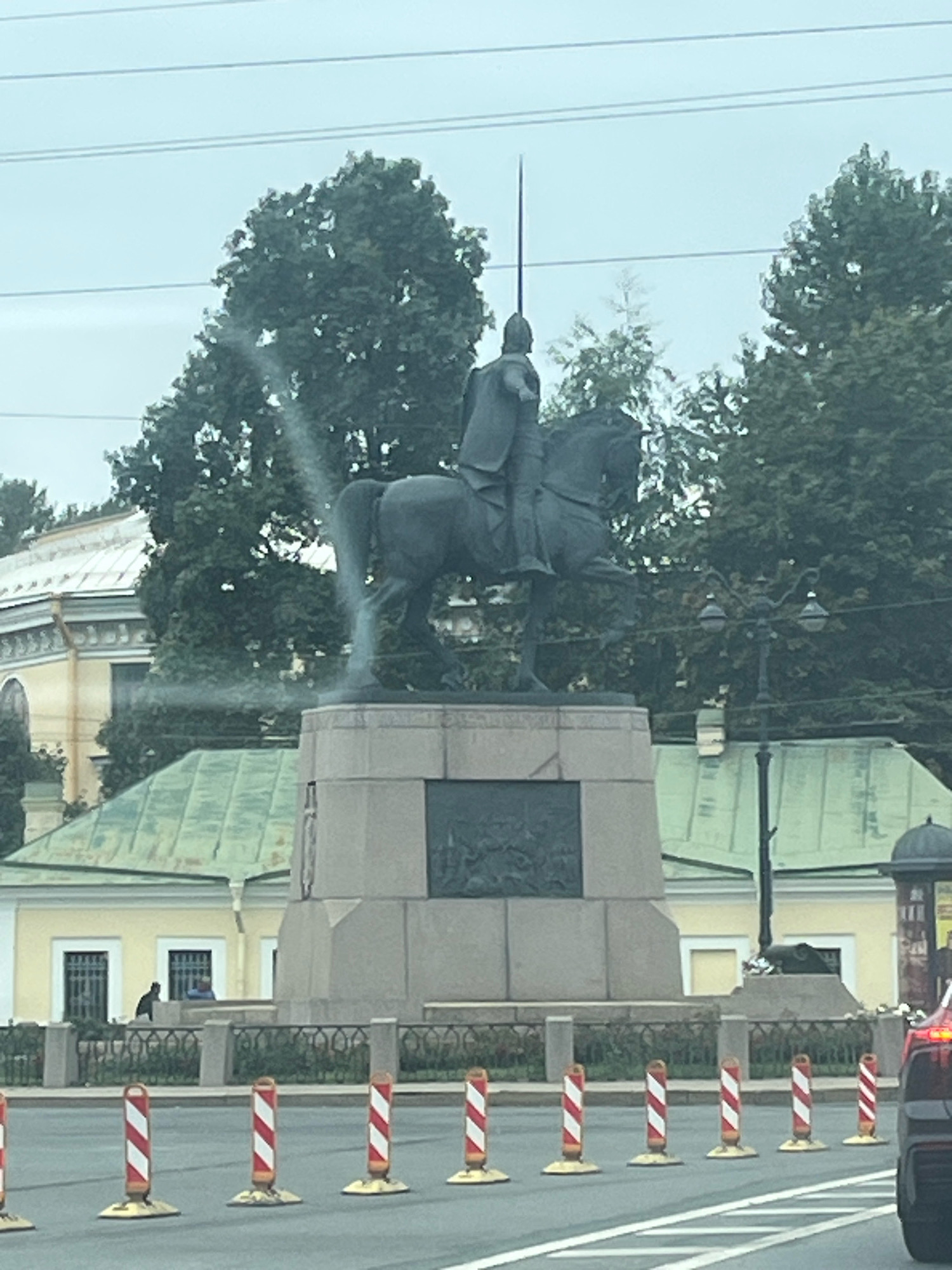 The image captures the Bronze Horseman statue, a famous landmark in Saint Petersburg, Russia, situated on Senate Square. The equestrian statue, depicting Peter the Great, stands atop a large granite pedestal. In the background, several trees with green foliage are visible, and beyond them, a pale blue sky with visible power lines.
To the left of the statue, a portion of a light yellow building with white trim and arched windows is visible. In front of this building, a low decorative fence runs along the edge of a walkway. A dark-colored figure, possibly a person, is seen near a window of the building.
The foreground of the image shows a street with a lane divider marked by white lines. Several temporary traffic barriers, cylindrical with red and white diagonal stripes, are placed along the edge of the road. A portion of a dark-colored vehicle is visible on the right side of the frame. The overall lighting suggests an overcast or cloudy day.