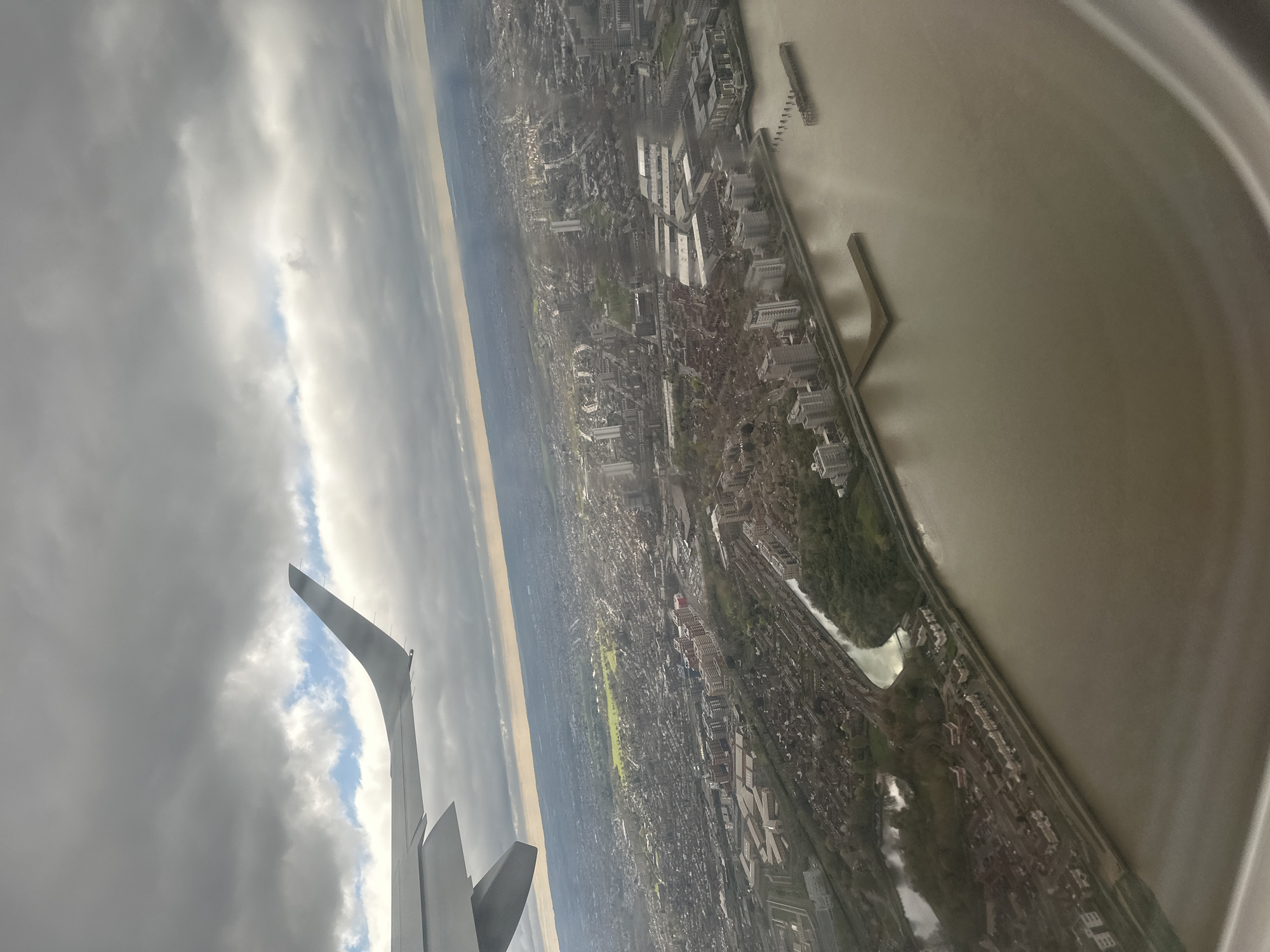 An aerial view from an airplane window captures a cityscape along a wide river, possibly the Thames, and part of the aircraft wing. The river's color is murky brown-grey. The city is dense with buildings of varying heights and styles. The sky is cloudy with grey tones, but there is a strip of lighter color. Some man-made structures jut into the river, like small docks.