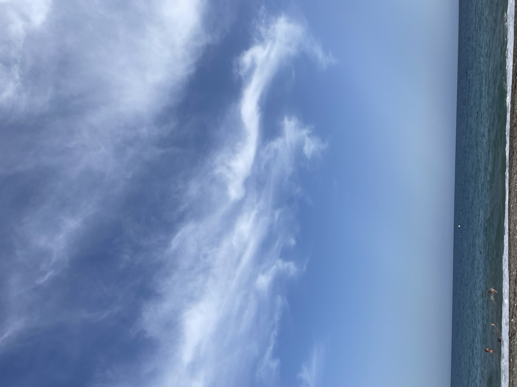 The image depicts a scenic view of the Mediterranean Sea from a beach in the municipality of Lújar, Spain. The sky above is a vibrant blue, adorned with wispy white cirrus clouds that streak across the upper portion of the frame. The sea is a deep blue, with gentle waves breaking on the pebbled shore. In the water, several individuals are visible, engaged in swimming or playing. One person is lying on their back, while others are partially submerged, with their heads and upper bodies visible. The beach itself is composed of small, smooth pebbles, typical of the region. The overall impression is one of a serene and inviting coastal environment.