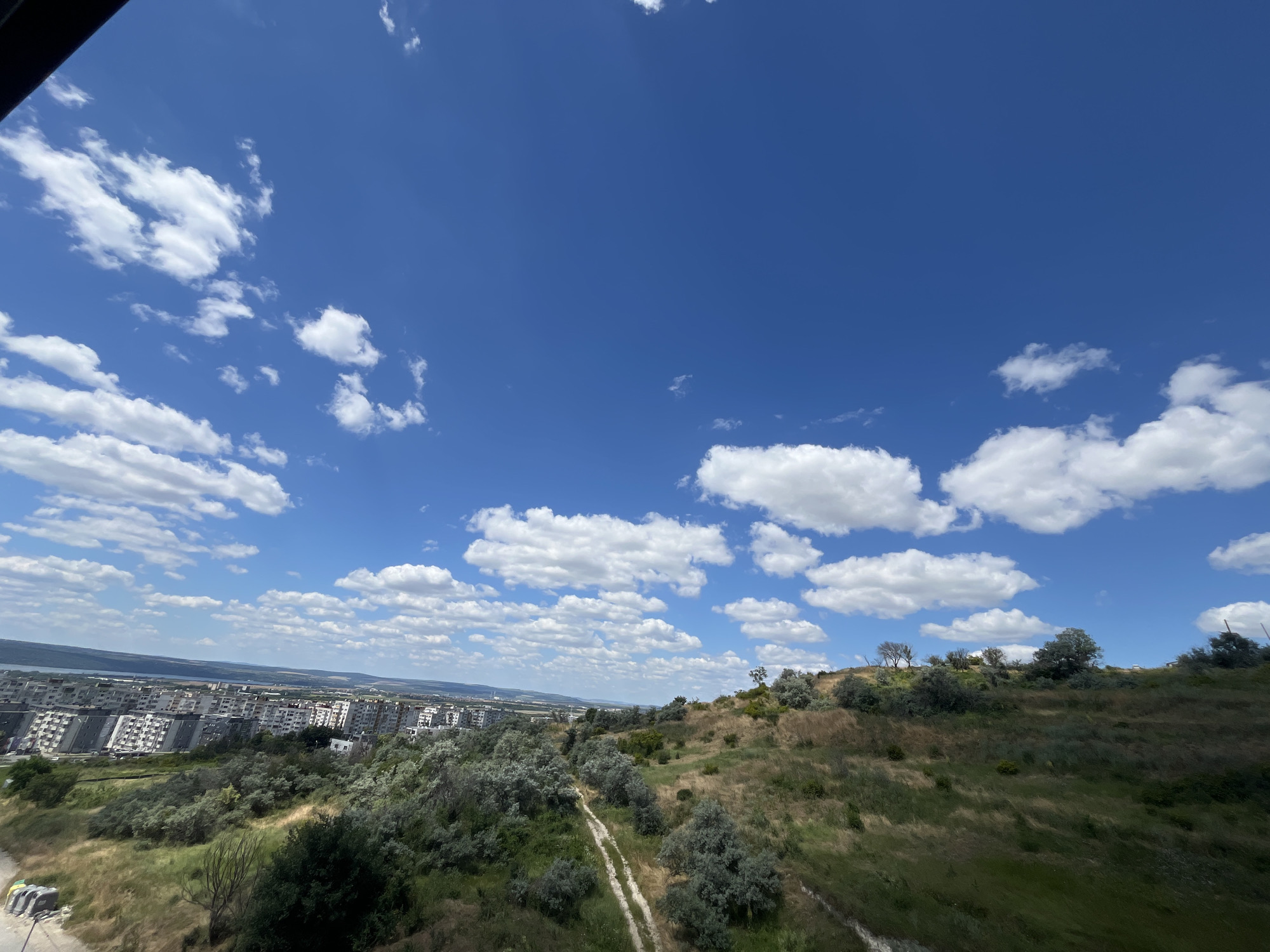 This is an eye-level, wide shot captured on a sunny day, featuring a vast expanse of blue sky dotted with fluffy white clouds. The foreground is dominated by a grassy hillside covered in dry grass and scattered trees. A dirt path winds its way up the slope. In the middle ground, a dense cluster of trees and shrubs obscures a portion of a city. The background reveals a cityscape with numerous apartment buildings, some appearing to be older Soviet-style architecture, stretching towards a body of water. The scene is consistent with a view from a hillside overlooking the Asparuhovo district of Varna, Bulgaria, likely in the vicinity of the Asparuhovo bridge or the park areas near it, looking towards the city center and the Varna Bay. No people or animals are visible in the image.
