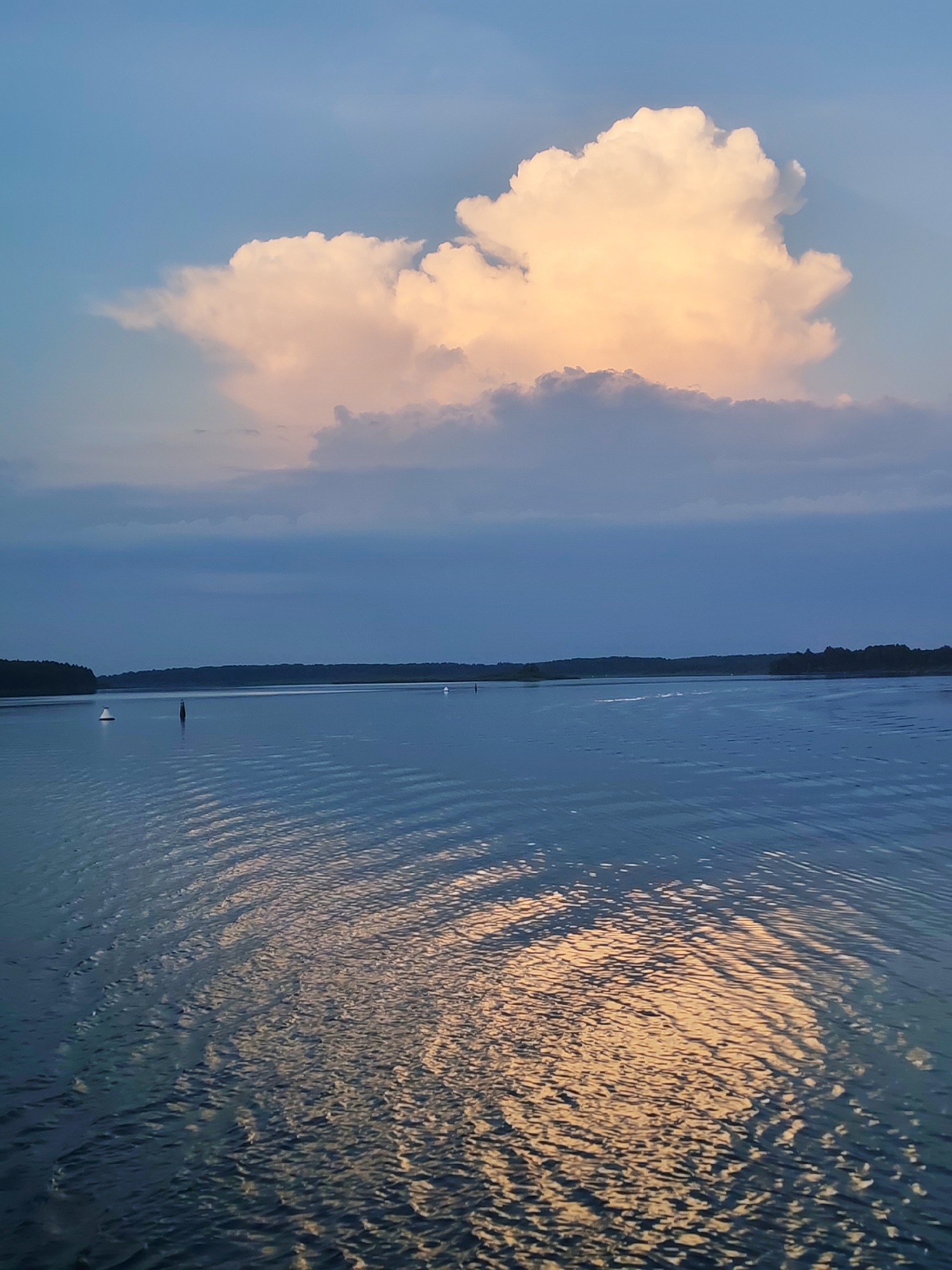 The image depicts a serene body of water under a sky with dramatic clouds. A large, fluffy cloud illuminated by warm sunlight dominates the upper portion of the frame, contrasting with the deeper blues of the surrounding sky and other, darker clouds. The water, rippling with the effect of movement or wind, reflects the light of the sky and the prominent cloud. Along the horizon, a dense line of trees forms a dark silhouette, suggesting a distant shoreline. Several buoys are visible in the water, indicating navigation markers. The overall impression is one of a peaceful, natural landscape during a time of day when light and shadow play across the scene. The location is identified as Edoma, Russia.