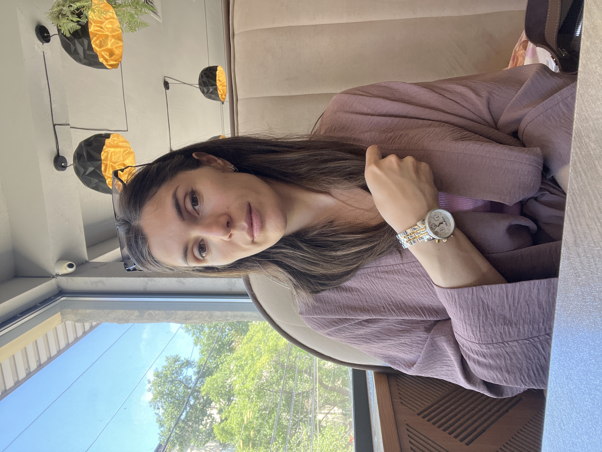A woman with long, dark brown hair is seated indoors, looking directly at the viewer with a neutral expression. She is wearing a dusty rose or light purple long-sleeved top made of a textured, crinkled fabric. A silver and gold-toned wristwatch with a white dial and chronographs is visible on her left wrist. Her left hand is resting, while her right hand is positioned near her shoulder. Glasses are perched on top of her head.
The setting appears to be a cafe or restaurant, featuring plush, light brown upholstered booth seating. The wall behind her is decorated with several black, geometric bowl-shaped fixtures that have golden interiors; some of these contain green foliage, resembling modern wall sconces or planters. A large window to her left reveals a bright, clear blue sky and lush green trees outside, indicating a sunny daytime. A small white security camera is mounted above the window. A dark wooden surface with a geometric pattern is visible in the foreground, likely part of a table or chair. No visible text is present in the image.