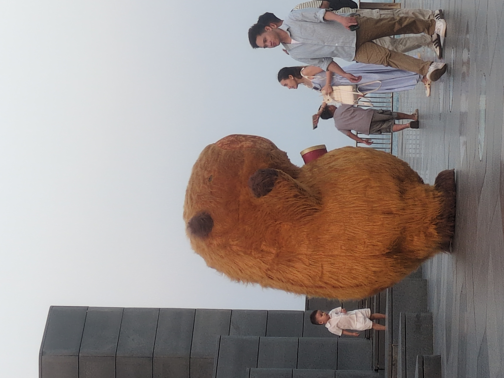 In Bình Kiến, Vietnam, a giant brown teddy bear sculpture sits on a platform accessible by gray concrete steps. A young child in white stands on the steps. Above the bear and to the side, several people are walking. One man in a striped shirt and khaki pants walks with a woman in a light blue dress and white bag. Another man follows, holding up a phone. The background is a hazy sky.