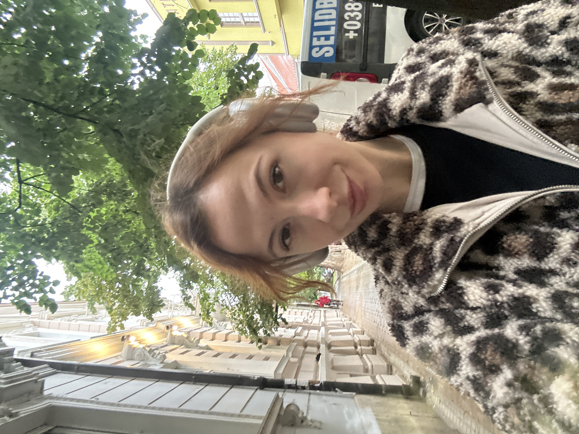A selfie portrait shows a fair-skinned woman with reddish-brown hair and light gray over-ear headphones, wearing a black top under a brown and white spotted, zip-up jacket. The photo is taken outdoors on a street in Novi Sad, Serbia. Behind the woman, trees overhang a sidewalk and street lined with light-colored buildings. A utility vehicle with "SELIDBE +038" visible on the side is also in the background. A cyclist is further down the street.