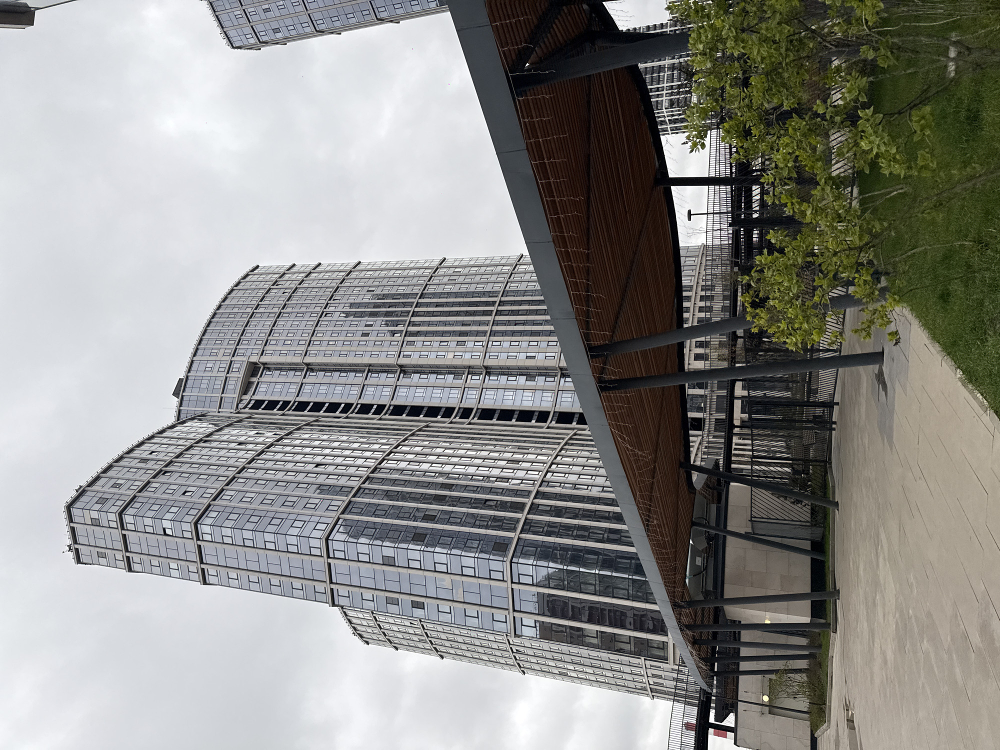 The image presents a modern urban landscape in Khimki, Russia, centered around a tall, contemporary building under an overcast sky. The primary subject is a multi-story high-rise with a distinctive curved glass facade, reflecting the cloudy conditions. Its exterior is characterized by numerous rectangular windows, creating a repeating pattern across its surface.
In the foreground and mid-ground, a long, covered walkway or pergola stretches horizontally across the scene. This structure features a slatted wooden ceiling supported by dark metal pillars. Adjacent to the walkway on the right, there is a manicured area of vibrant green grass with several small, leafy trees and bushes, indicating a landscaped public space. A light-colored paved path runs alongside the greenery and beneath the covered structure. A dark metal fence is visible behind the pergola, segmenting the area.
The weather is overcast with a uniformly grey sky, indicating a cloudy daytime setting without direct sunlight. There are no people visible, and no specific activity or event is taking place, contributing to a tranquil and still depiction of a well-maintained urban environment. No legible text is visible within the frame.