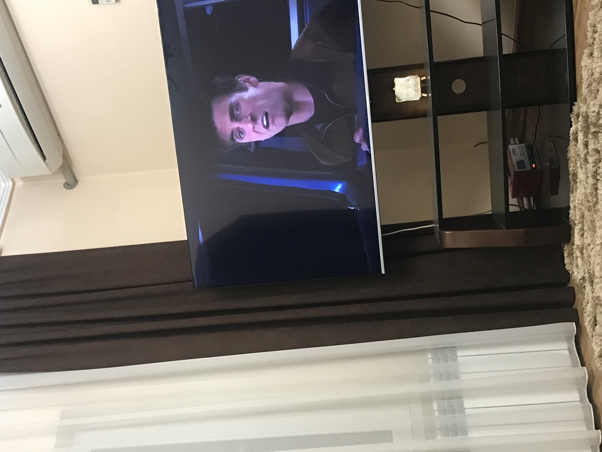 The image shows a television screen displaying a man looking surprised. The TV is positioned in front of a dark brown horizontal panel or curtain, which is in turn above a white window blind. To the right of the TV is a dark-colored shelving unit, holding various electronic devices. A beige carpet is also visible on the right side of the shelving unit. The location is not discernible from the image.