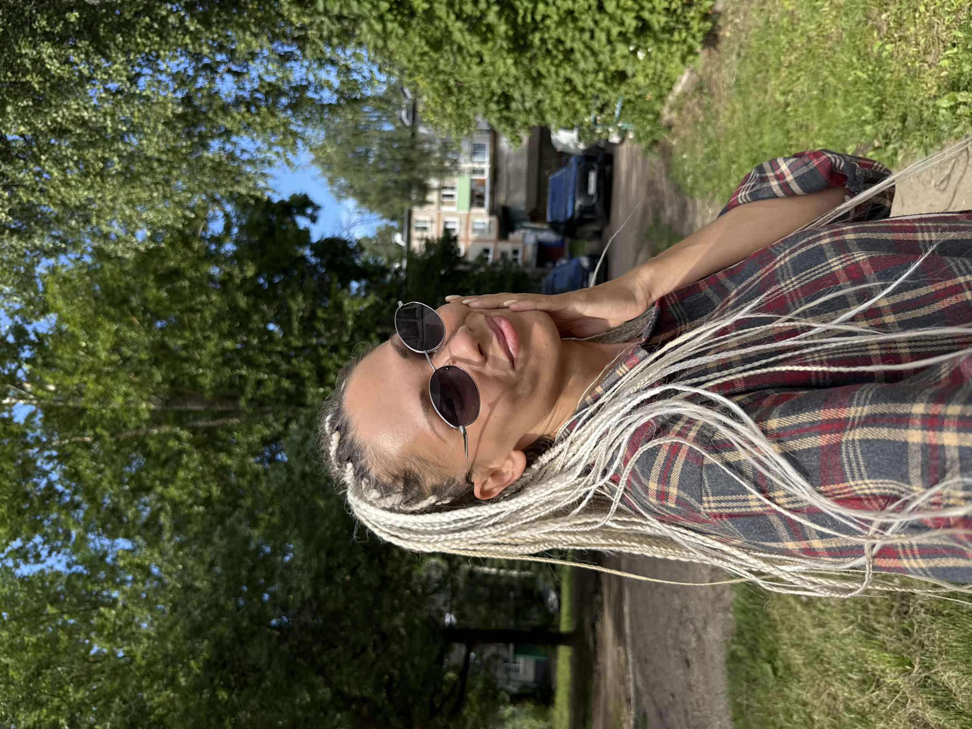 A woman with light blonde braided hair is captured in a selfie outdoors. She is wearing a plaid shirt and sunglasses, and her hand is resting on her cheek. The background features trees, a building, and parked cars, suggesting an urban or suburban environment. The image was taken in Rusanovo, Russia.