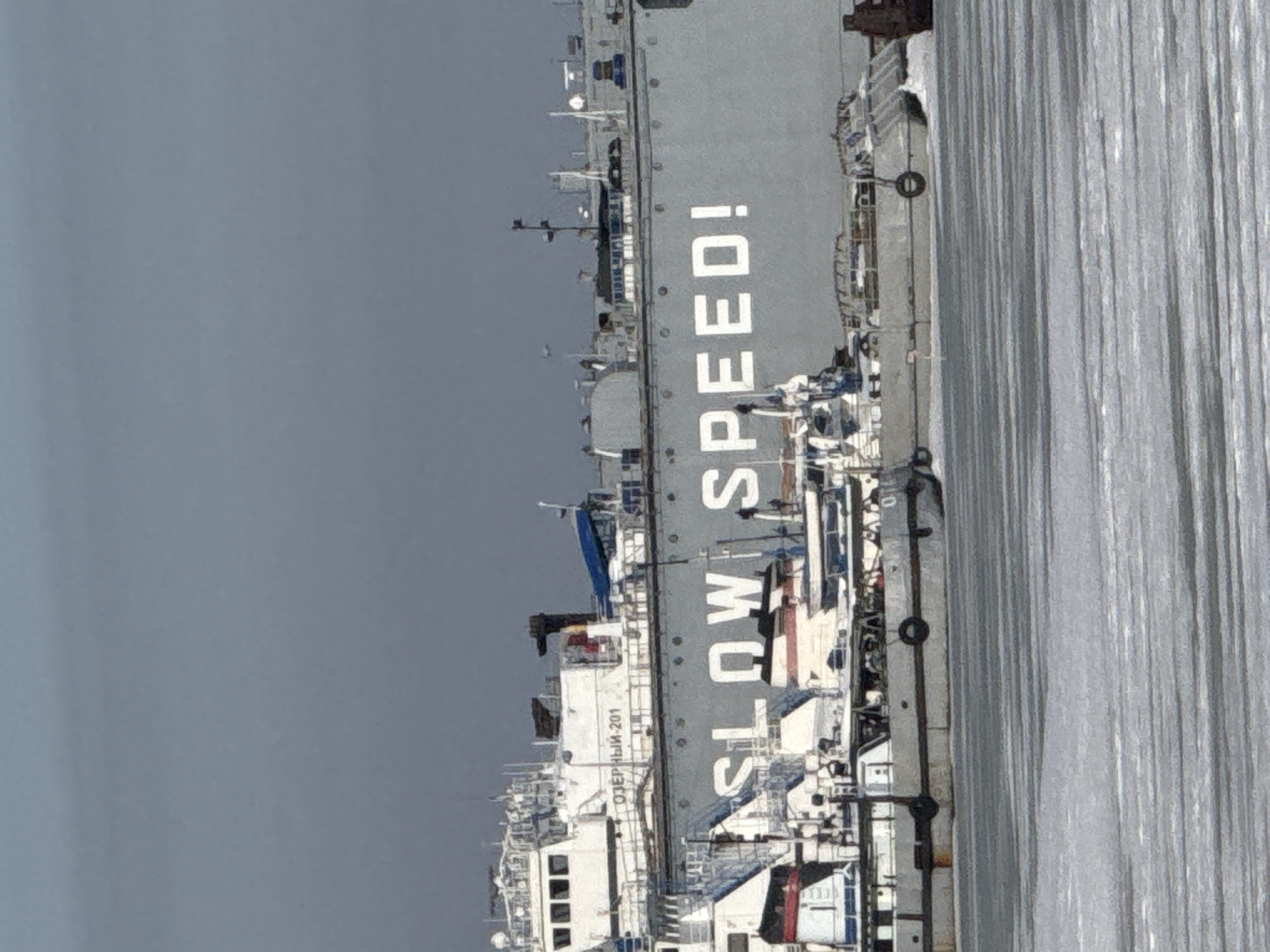 The scene depicts a frozen industrial waterway in the city of Chaikovsky, Russia, under an overcast grey sky. A large, grey cargo ship, identified by the white Cyrillic text "ОЗЕРНЫЙ-201" on its upper hull and the prominent English warning "SLOW SPEED!" painted in large white letters across its side, occupies the midground. The ship features a multi-deck white superstructure with windows, radar, and antennae on its port side. Several smaller workboats, painted in various shades of grey and white, are moored in the foreground, partially obscuring the lower hull of the larger vessel. One smaller vessel has black tire fenders affixed to its side. The surface of the water in the immediate foreground is covered in patterned sheets of ice, consistent with cold weather conditions. No human figures are visible.
