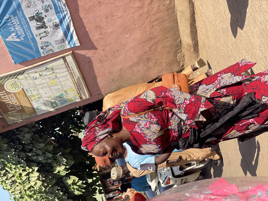 The image depicts a bustling outdoor scene in Samaru Ultra Modern Market, Samaru, Zaria, Kaduna State, Nigeria. In the foreground, a woman with dark skin wears a vibrant red dress with a yellow, white, and black leaf-like pattern, a matching headtie, and large hoop earrings. She carries a brown leather shoulder bag with a gold 'B' emblem and has a black fabric wrapped around her waist. She is smiling and looking to her right.
Behind her, several individuals are visible: a man in a light brown traditional garment and a dark cap, another man in a light blue t-shirt and a light brown woven cap, and other indistinct figures suggesting a crowd. A white and silver motorcycle is partially visible in the midground to the left.
The background on the right consists of a salmon-pink textured wall with two signs mounted. The left sign, framed in wood, advertises "Arsalan STORES" for "High Quality Skincare & Haircare Products." The right sign is a blue banner for "AL-MUBAARAK VARIETY MALL," offering "AFFORDABLE PHONE AND ACCESSORIES, HOUSEHOLD EQUIPMENT AND KITCHEN ITEMS," and explicitly states its location as "Samaru Ultra Modern Market... Samaru, Zaria, Kaduna State." Dense green tree foliage is visible in the upper left background. The scene is brightly illuminated by direct sunlight, casting shadows on the wall and ground, indicating daytime activity in a commercial area.