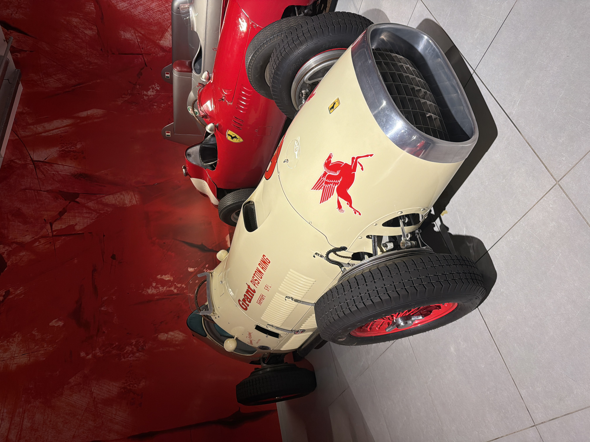 The image shows two vintage Formula 1 race cars displayed indoors, likely in a museum or exhibition. The car in the foreground is cream-colored with red detailing, including its wheels and a stylized winged horse logo on the hood. Text on the side reads "Grant PISTON RING Ferrari SPL". The car behind it is bright red with yellow accents and a prominent Ferrari emblem. Both cars are parked on a tiled floor in front of a textured red wall that appears to be a backdrop or a painted mural. The lighting suggests an indoor, controlled environment. Based on publicly available information, this scene is located at the Louwman Museum in The Hague, Netherlands, which houses a significant collection of classic cars. There are no people visible in the image.