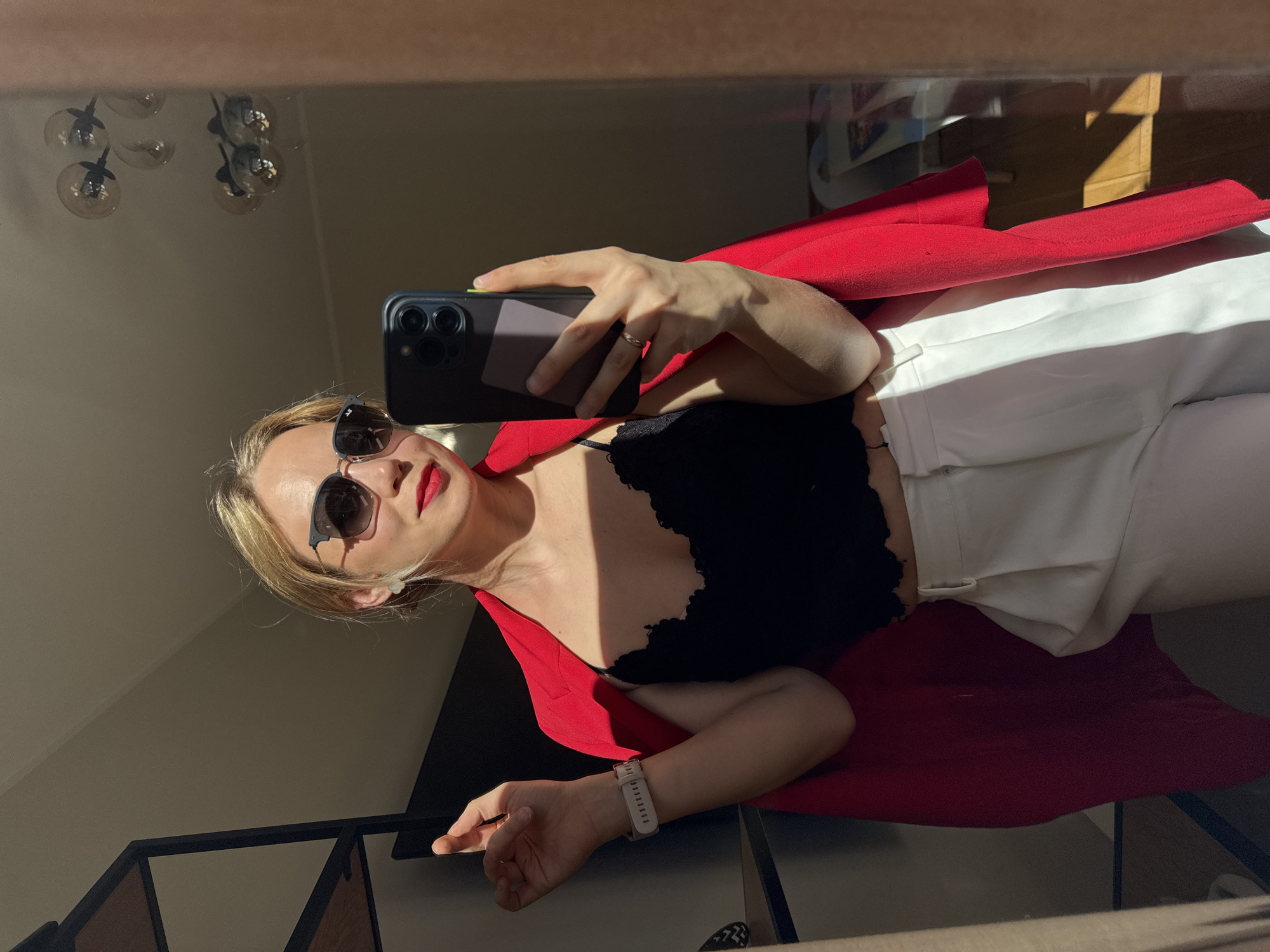 This image is a mirror selfie taken by a woman indoors, possibly in Riga, Latvia. She is captured from the chest up, angled slightly. She has blonde hair and wears dark sunglasses, which obscure her eyes, but her prominent red lipstick is clearly visible.
The woman is dressed in a vibrant red blazer, which is open to reveal a black lace crop top underneath. She also appears to be wearing white trousers. On her right hand, she holds a dark-colored smartphone with multiple rear cameras, positioned to take the photo. A simple, thin ring is visible on her right ring finger, and a light-colored smartwatch with a white band is worn on her left wrist.
The scene is illuminated by strong, direct sunlight, casting sharp shadows across her clothing and the background, suggesting either early morning or late afternoon. The background, visible in the mirror's reflection, includes a room with a light-colored wall and a distinctive chandelier with multiple glass globes hanging from the ceiling. A dark railing or shelving unit is also reflected in the lower portion of the image. The overall atmosphere conveys a stylish and well-lit indoor setting. No visible text is present in the image.