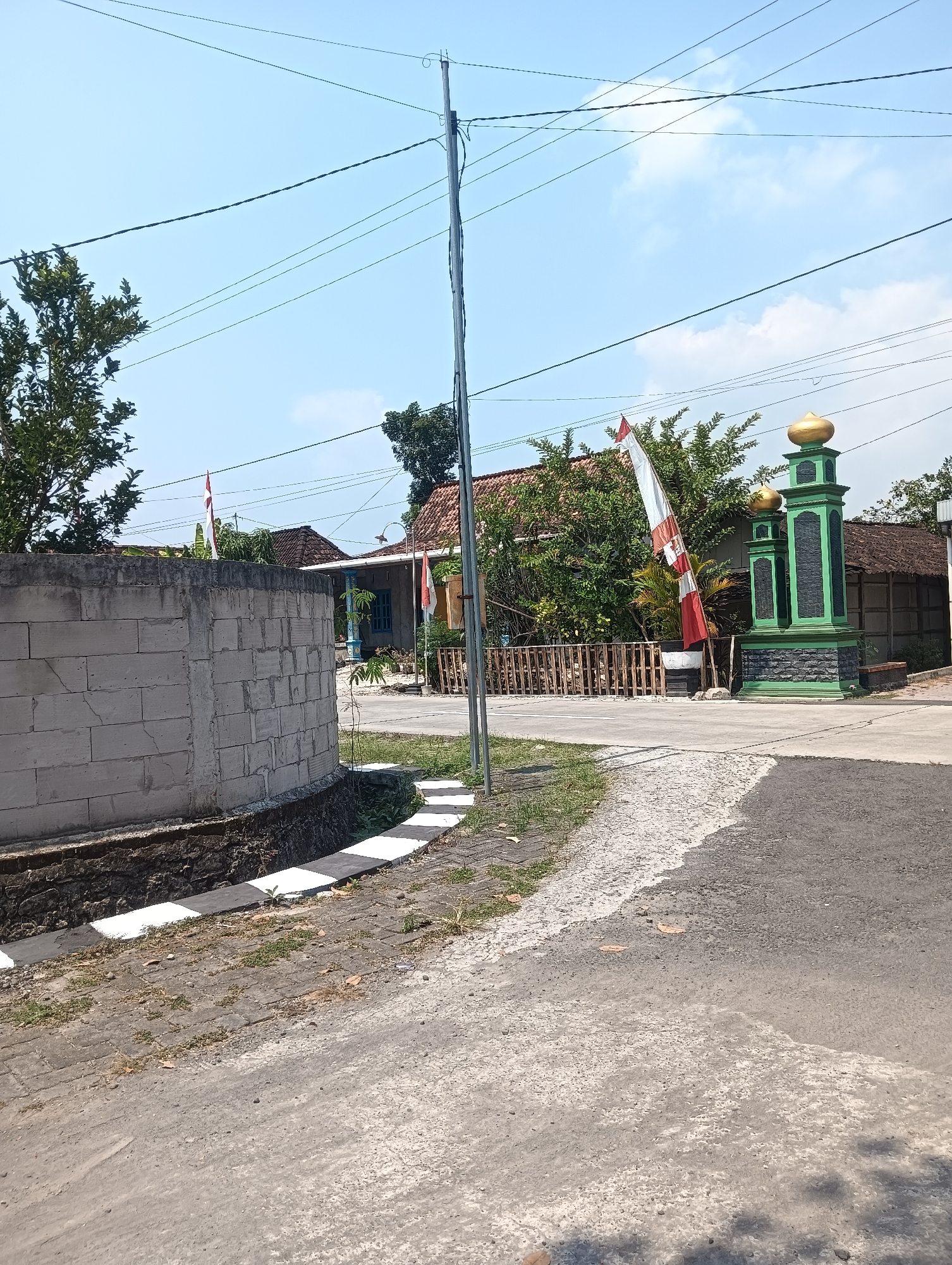 The image captures a street scene in Tanggan, Indonesia, under a bright, partially cloudy sky. In the foreground, a paved road transitions from asphalt to a lighter, textured surface on the left. Adjacent to the road on the left is a low, unfinished concrete block wall. Behind the wall, a drainage ditch with white and black striped concrete edges is visible.
Further into the scene, a wooden fence runs along the edge of the property. Two poles with Indonesian flags, red and white, are planted near the fence. To the right of the flags, a decorative green structure with two gold domes, resembling a mosque entrance or marker, stands prominently. Behind this structure and fence, houses with tiled roofs are visible, surrounded by trees and vegetation. Overhead, a network of utility wires crisscrosses the sky, supported by a tall metal pole on the right side of the frame. The overall impression is a typical residential street in a rural or semi-urban area of Indonesia.